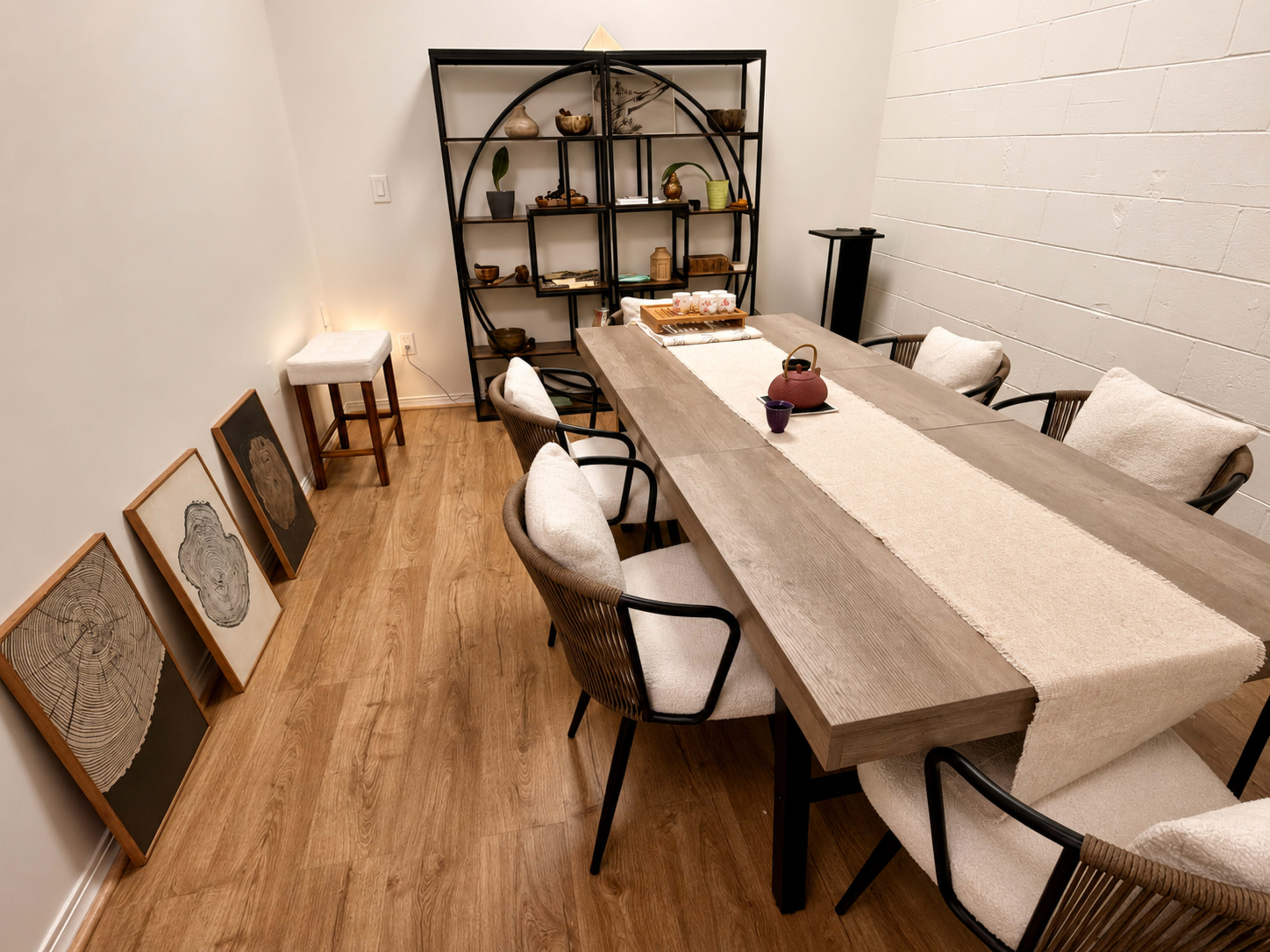 A minimalist dining room features a long wooden table surrounded by six light-colored chairs, with a decorative shelf displaying various objects against the wall.