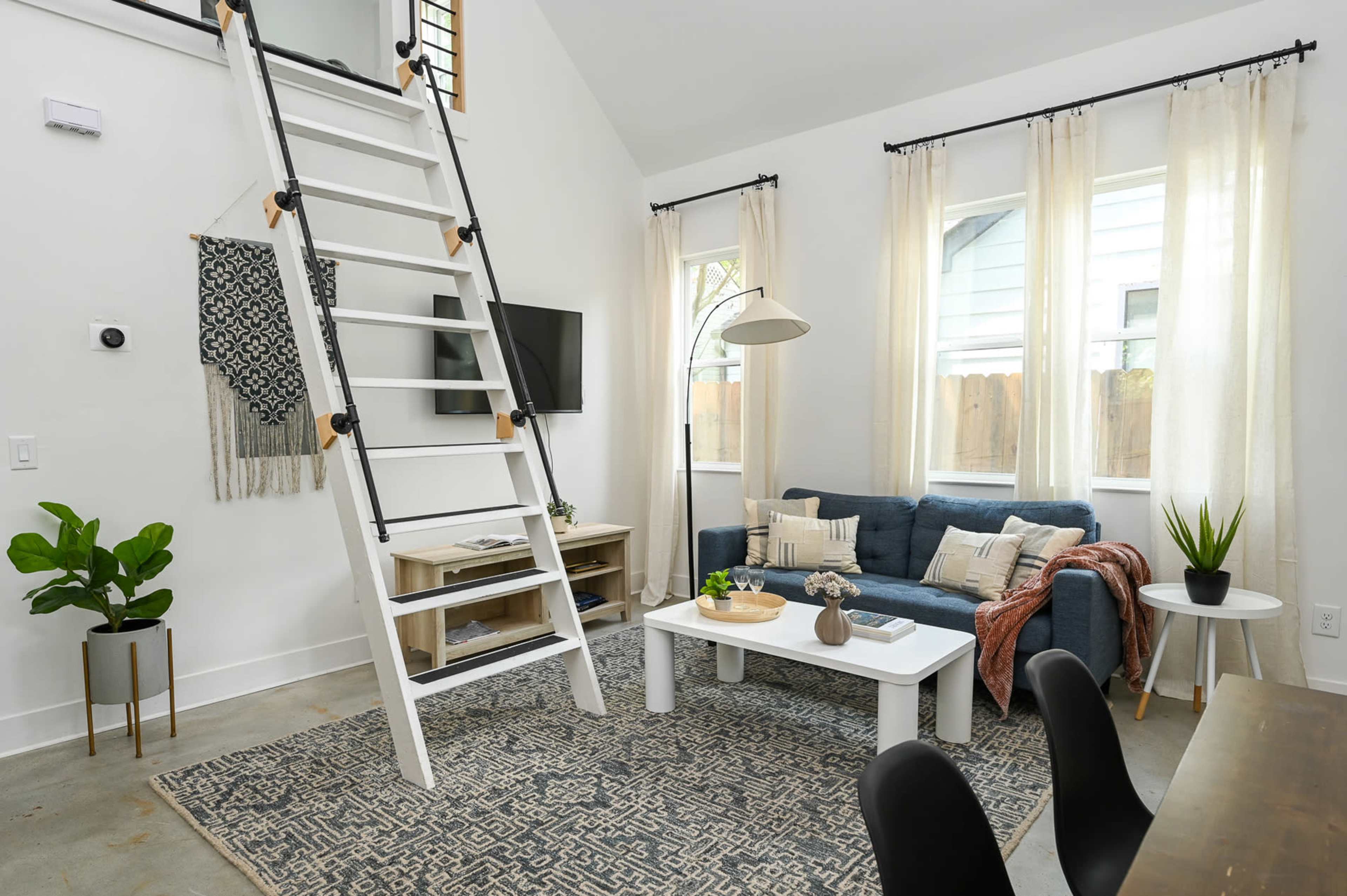 The image shows a modern living area featuring a blue sofa, a white coffee table, a decorative rug, and a wooden ladder leading to a loft space, with large windows providing natural light.
