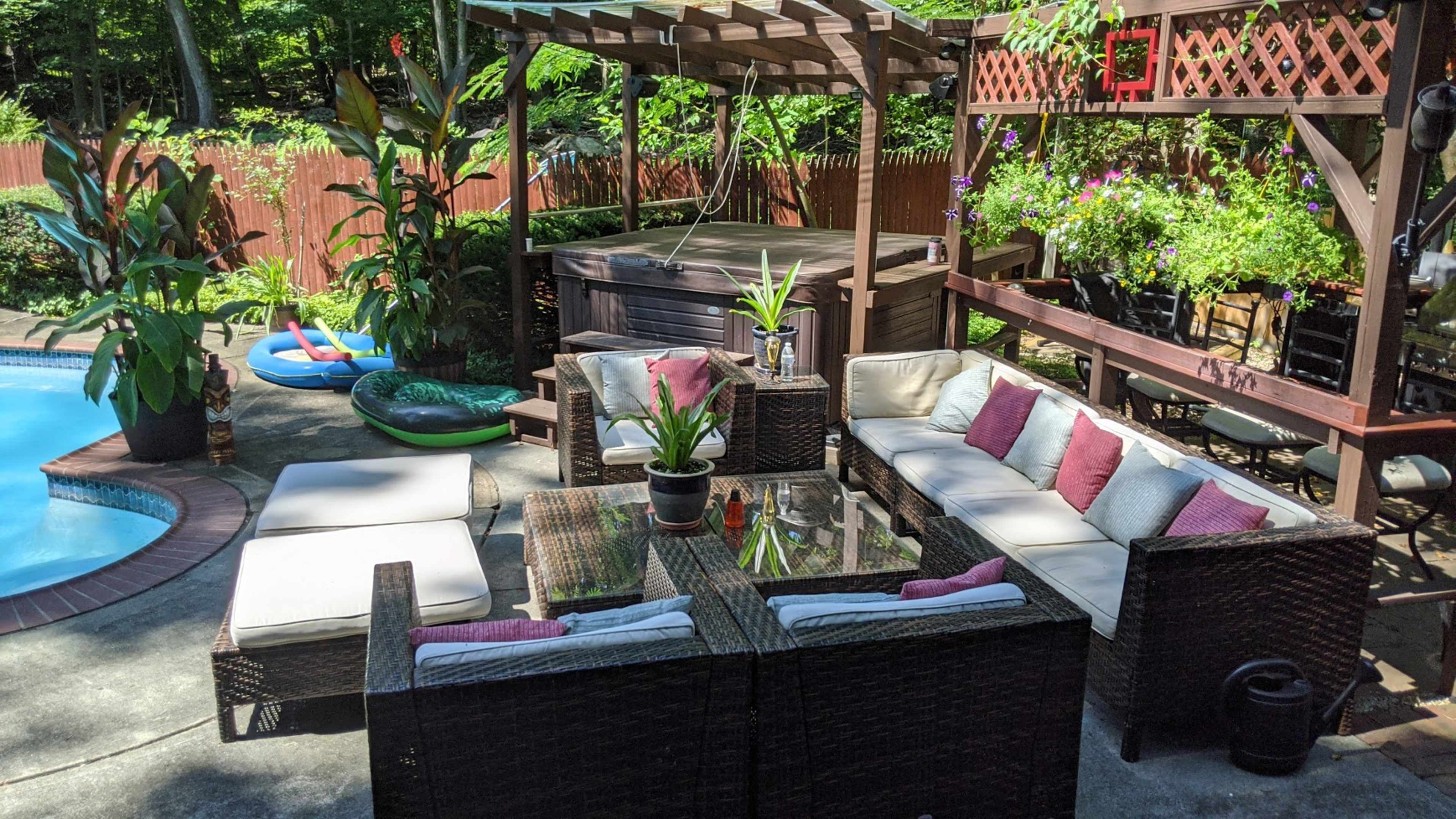 A patio area with a sectional wicker seating arrangement, a glass-top table, a hot tub, and various plants surrounding a swimming pool.