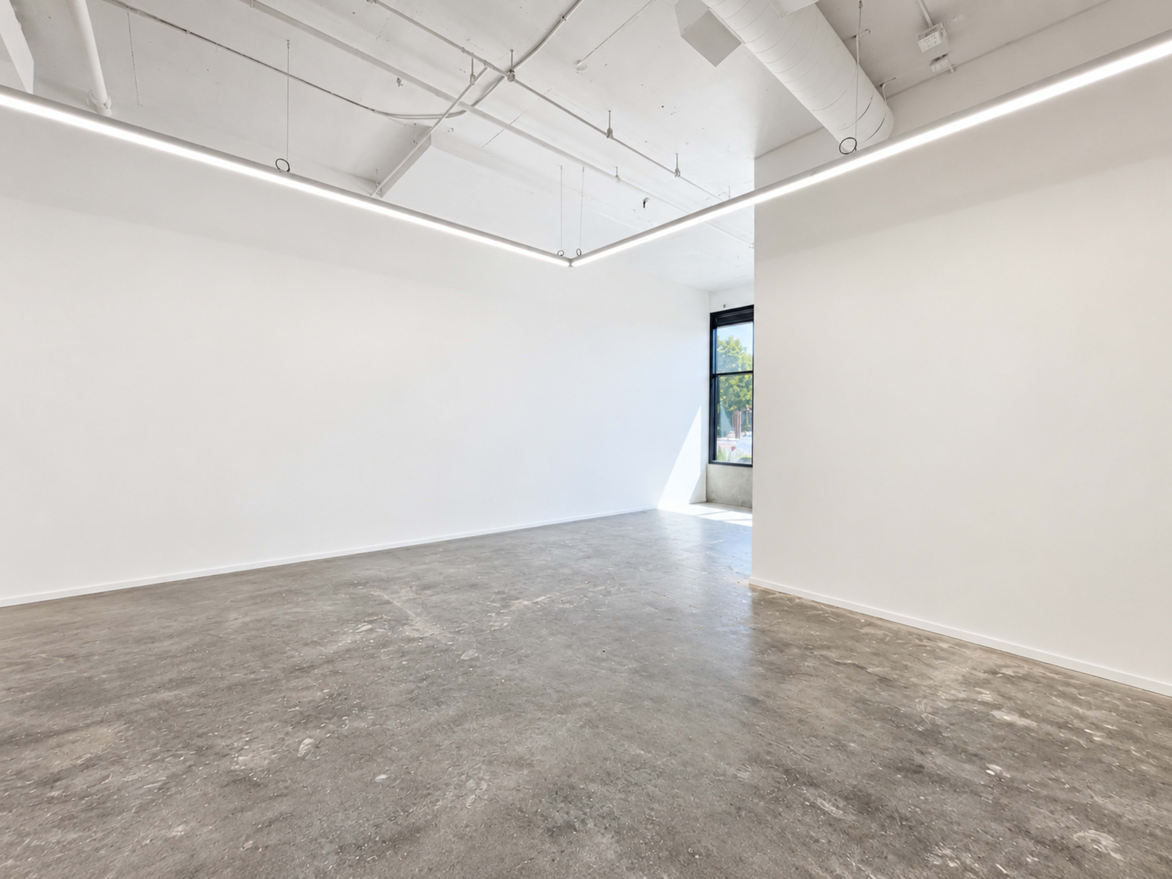 The image shows a spacious, empty room with concrete flooring, white walls, and a large window allowing natural light to enter.