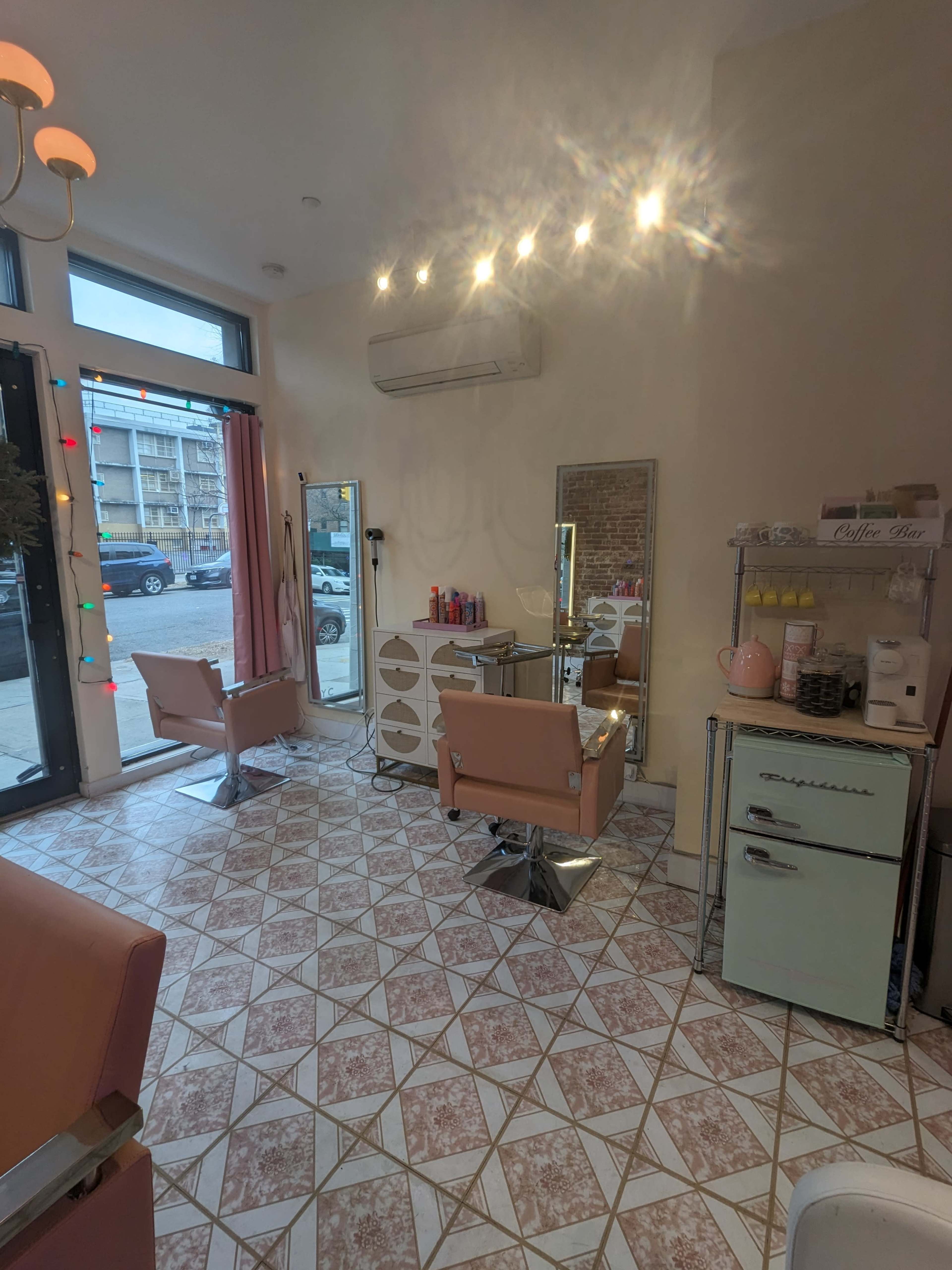 A vintage-styled hair salon with two pink chairs, large mirrors, and patterned tile flooring, illuminated by overhead lights.