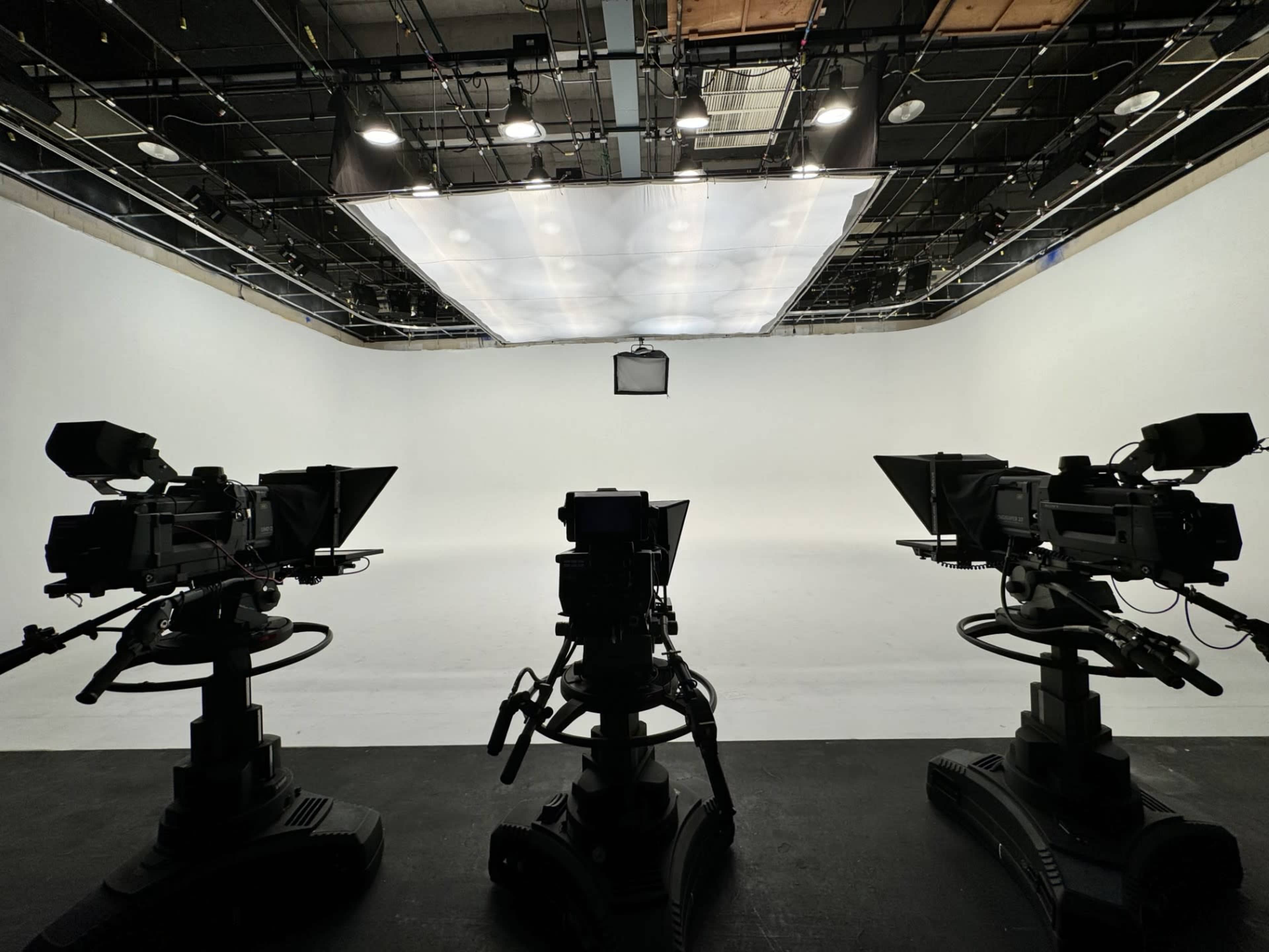 Three television cameras are positioned on tripods facing a blank white backdrop in a studio setting.