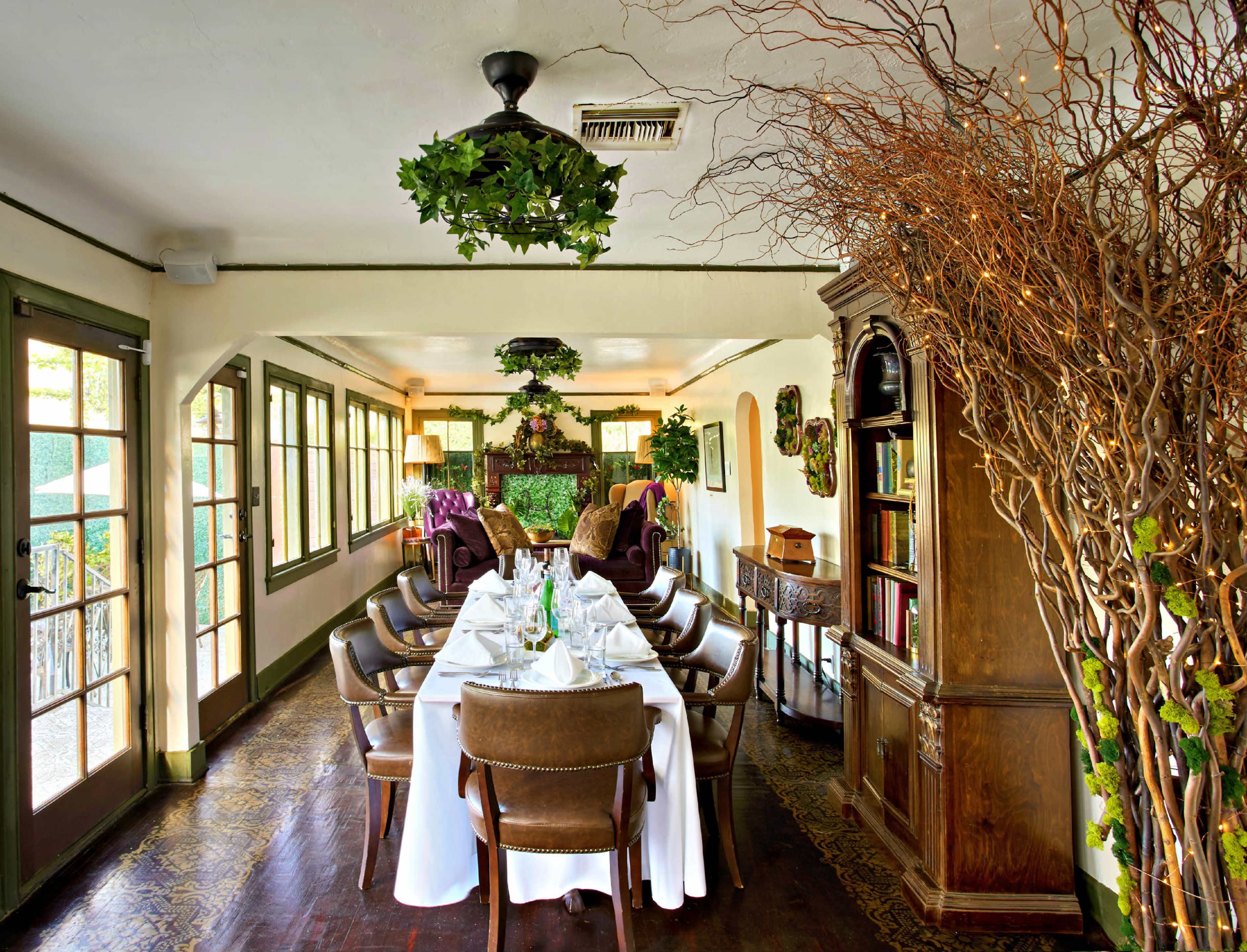 A long dining table is set with white tablecloths in a quaint room adorned with plants and wooden furniture.