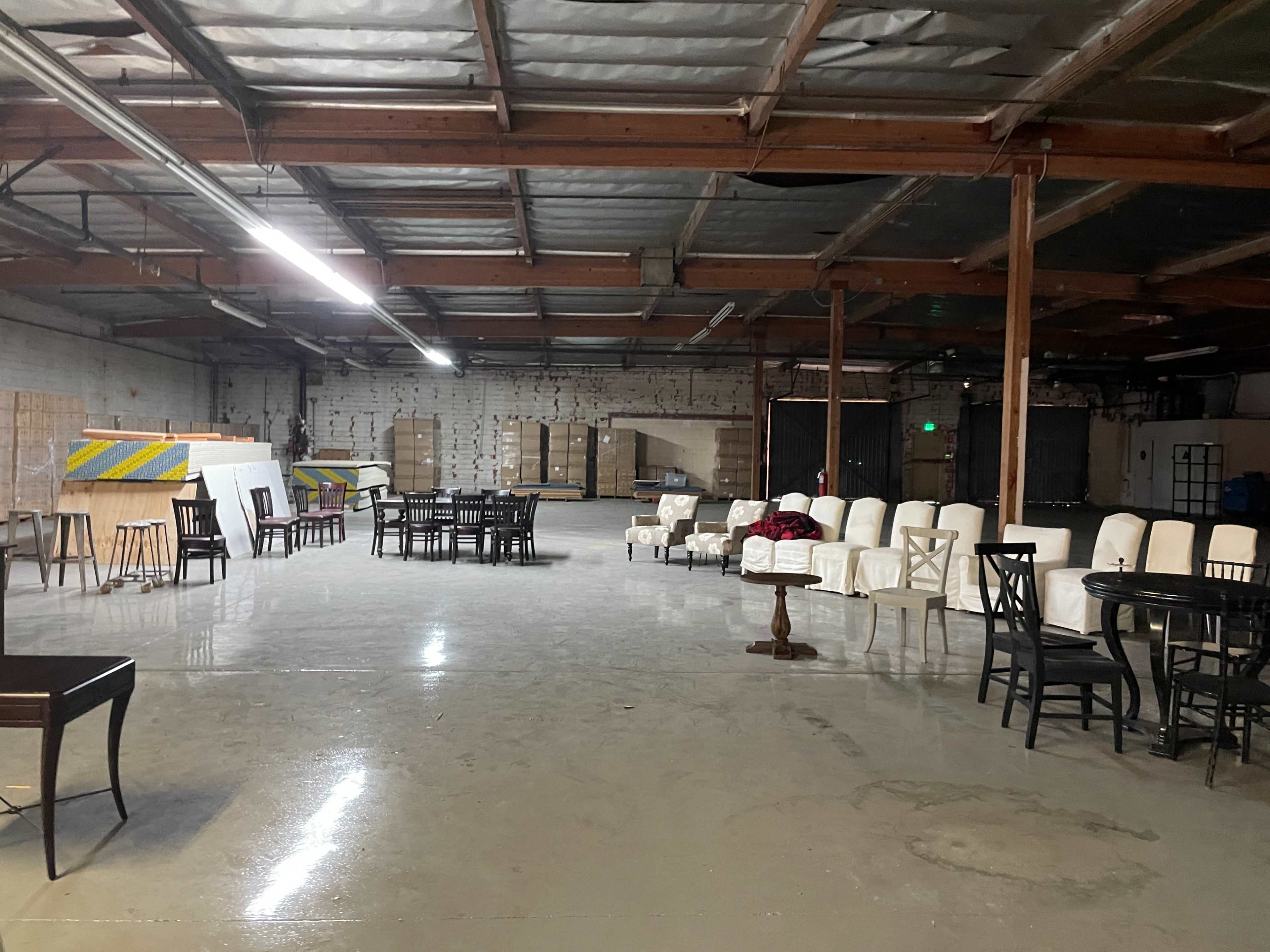 A spacious warehouse interior with various tables and chairs arranged throughout the area, surrounded by stacks of materials and plywood.