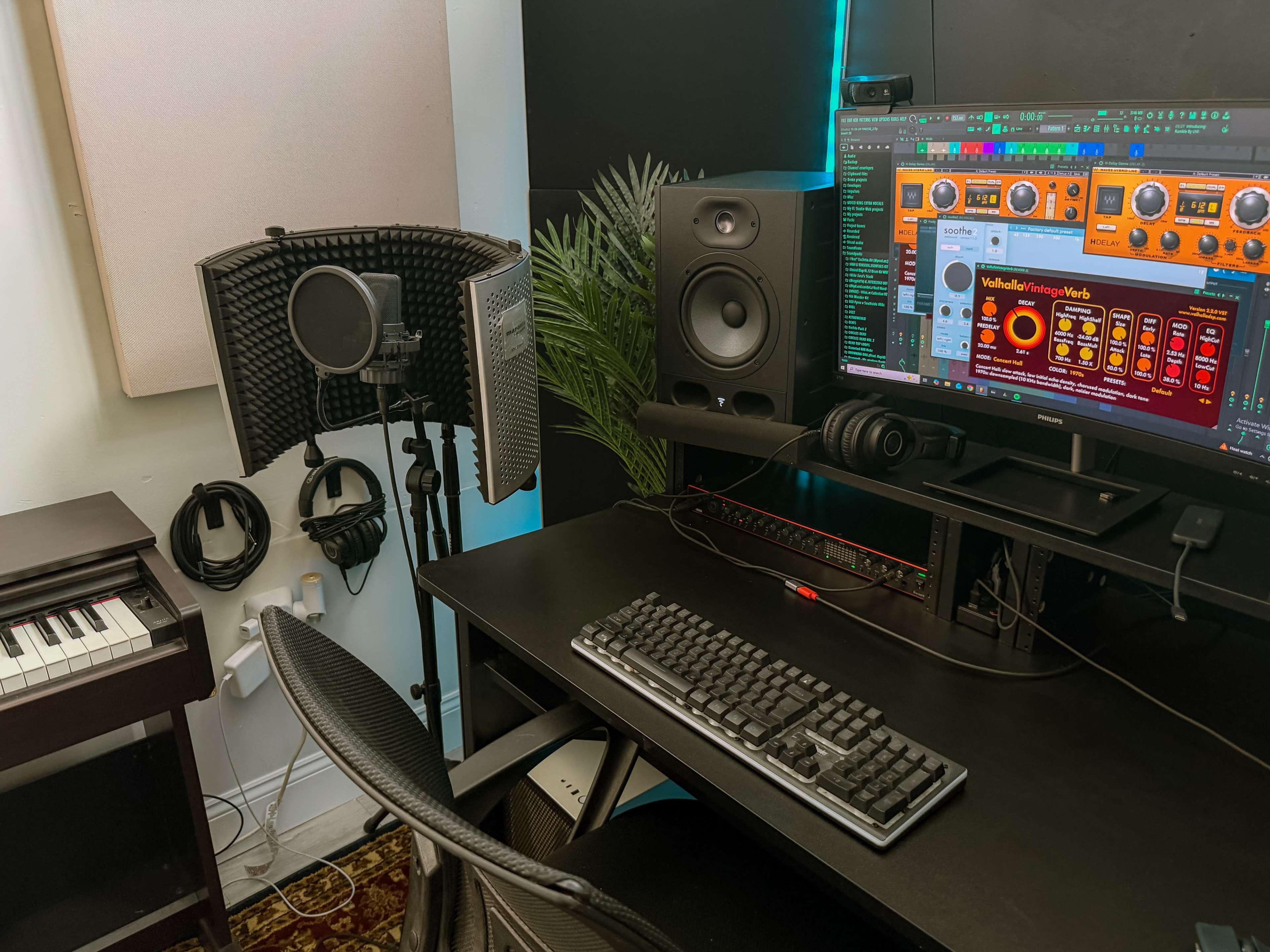 The image shows a compact music production studio featuring a computer desk with a keyboard, audio interface, studio monitors, and a microphone set up in a corner.
