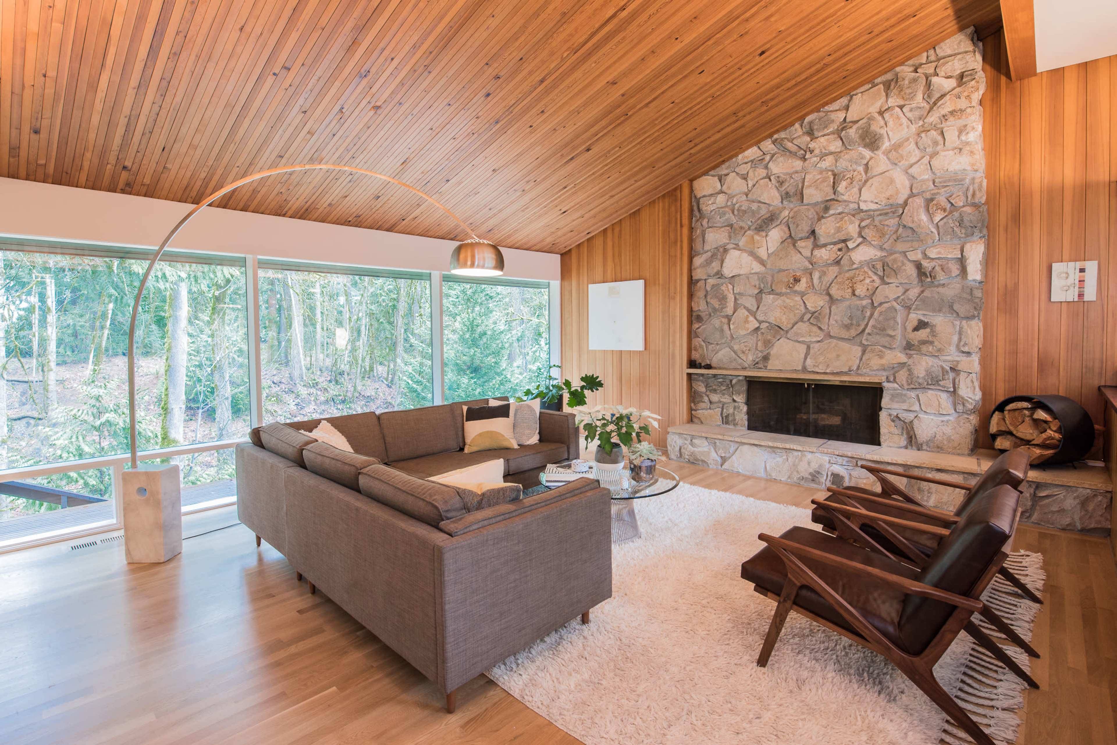 A modern living room features a stone fireplace, large windows overlooking greenery, and minimalist furniture including a gray sofa and wooden chairs.