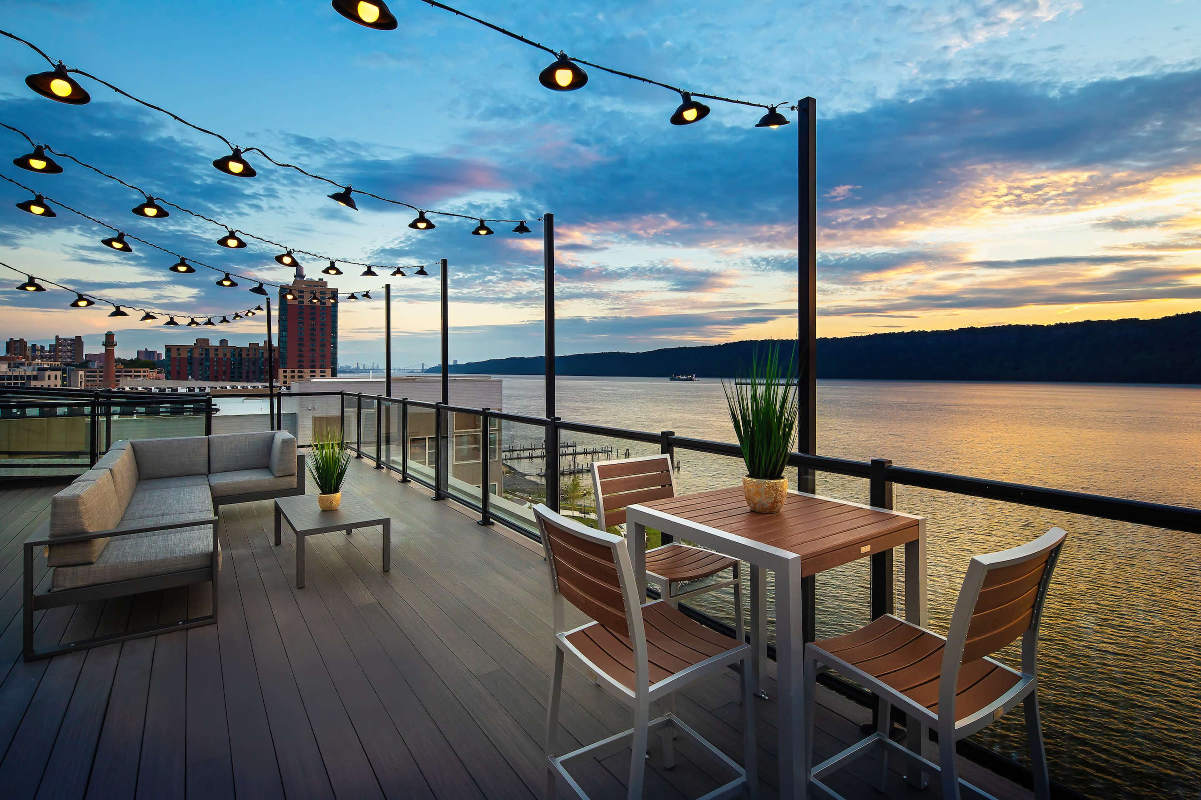 The image shows a modern outdoor balcony with string lights overlooking a river during sunset.