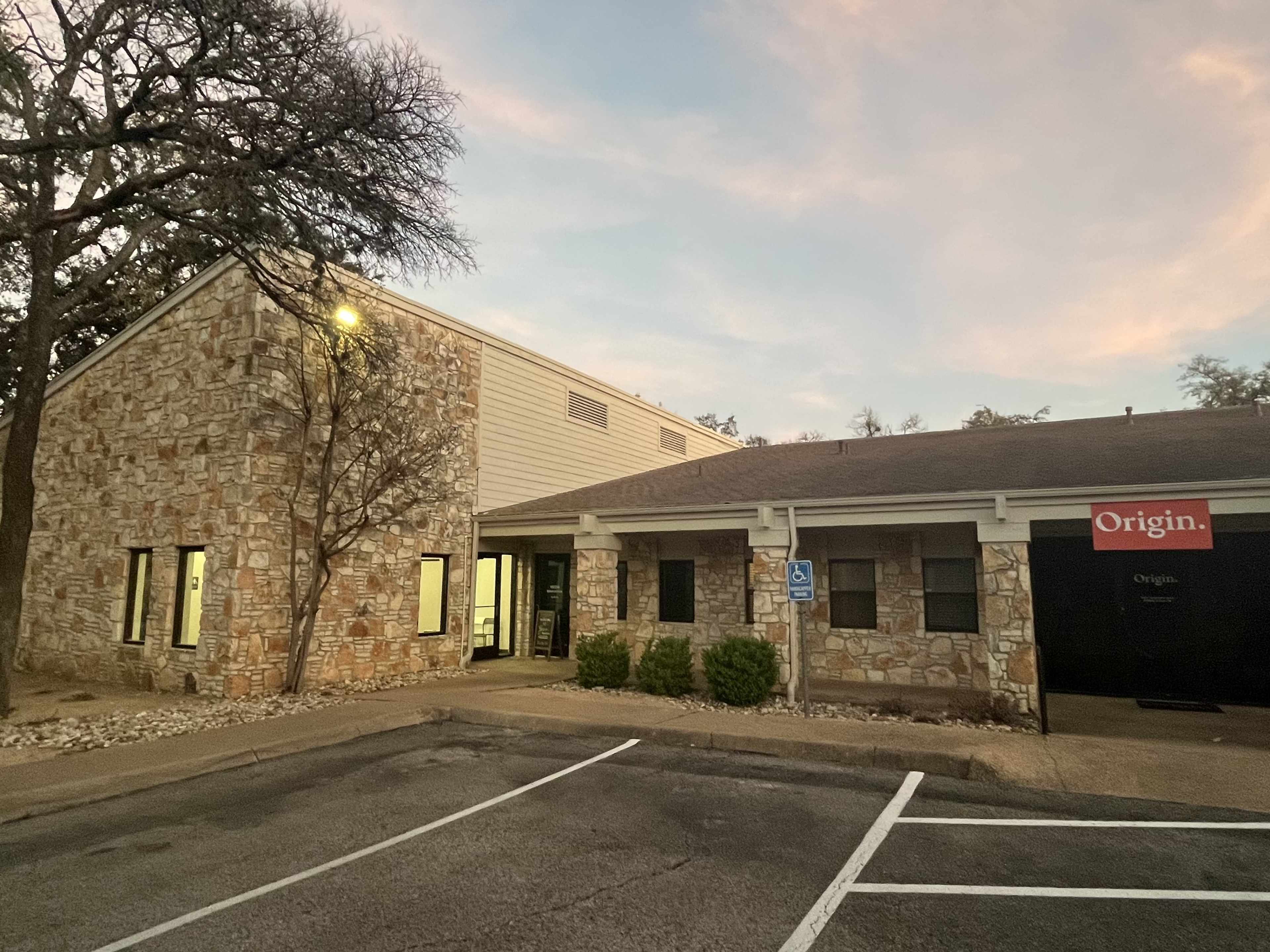 A building with a stone facade and a sign reading "Origin," located in a parking lot at dusk.