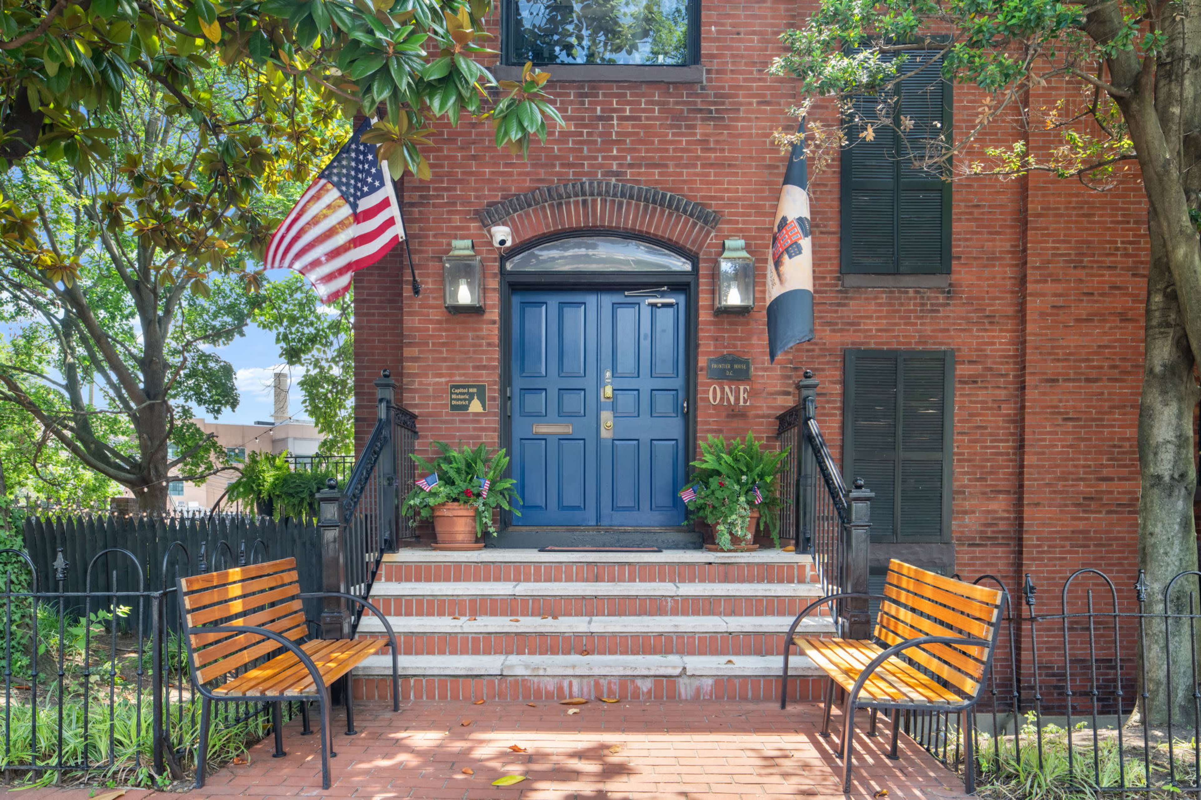 The image shows a red brick building with a blue front door, flanked by two flags and bordered by two wooden benches on a brick walkway.