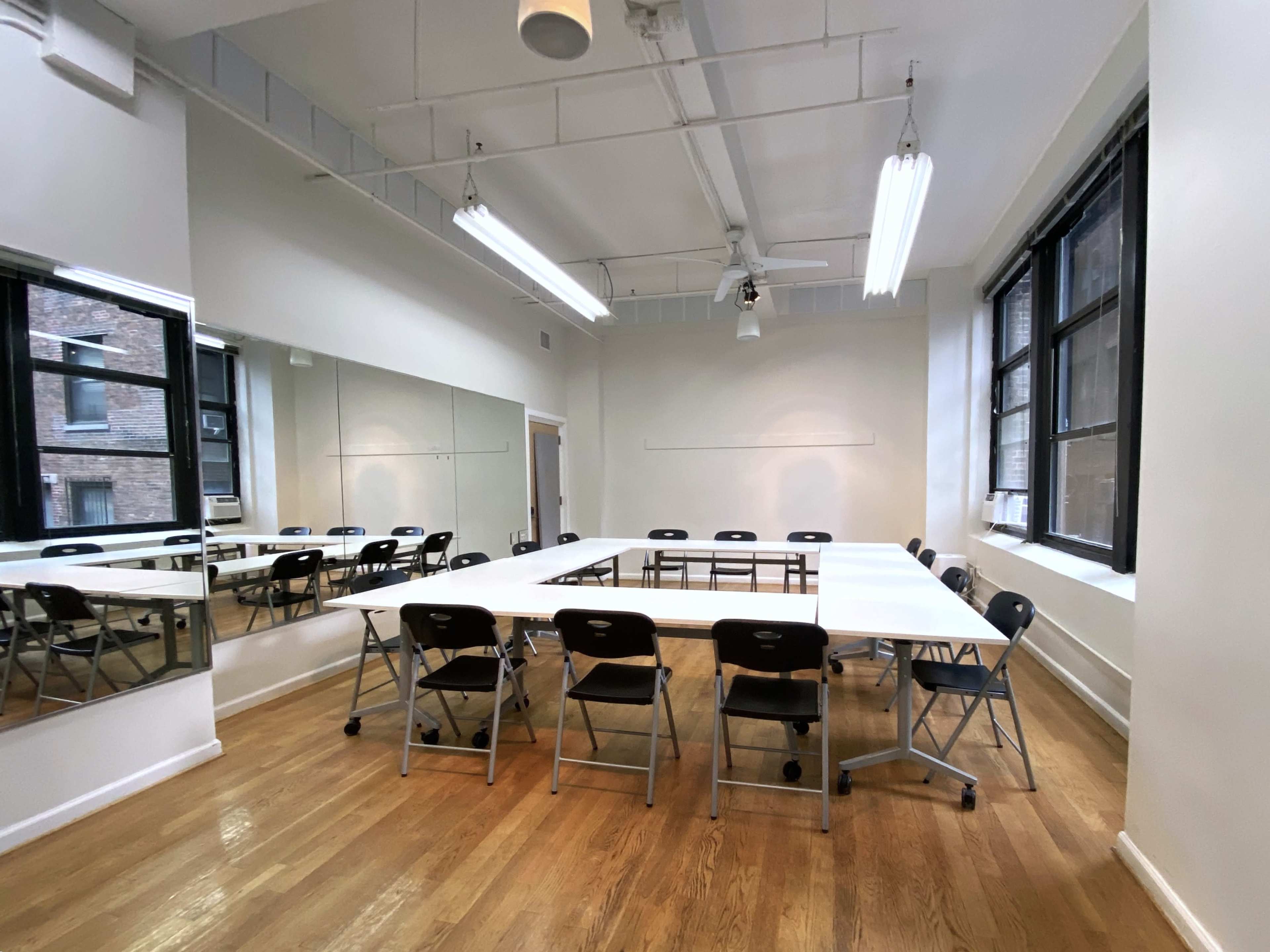 A bright, minimalistic meeting room features a large rectangular table surrounded by folding chairs, with windows and mirrors lining the walls.
