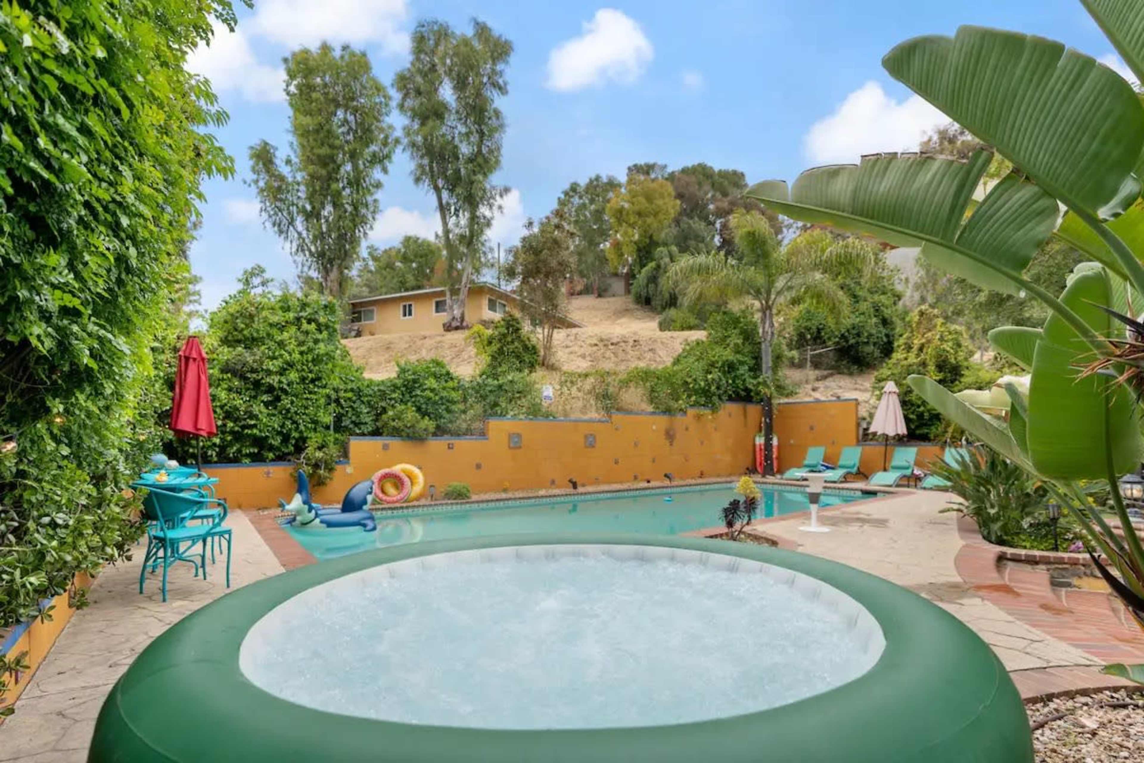 A backyard scene features a swimming pool surrounded by greenery and lounge chairs, with a hot tub on the side and umbrellas offering shade.