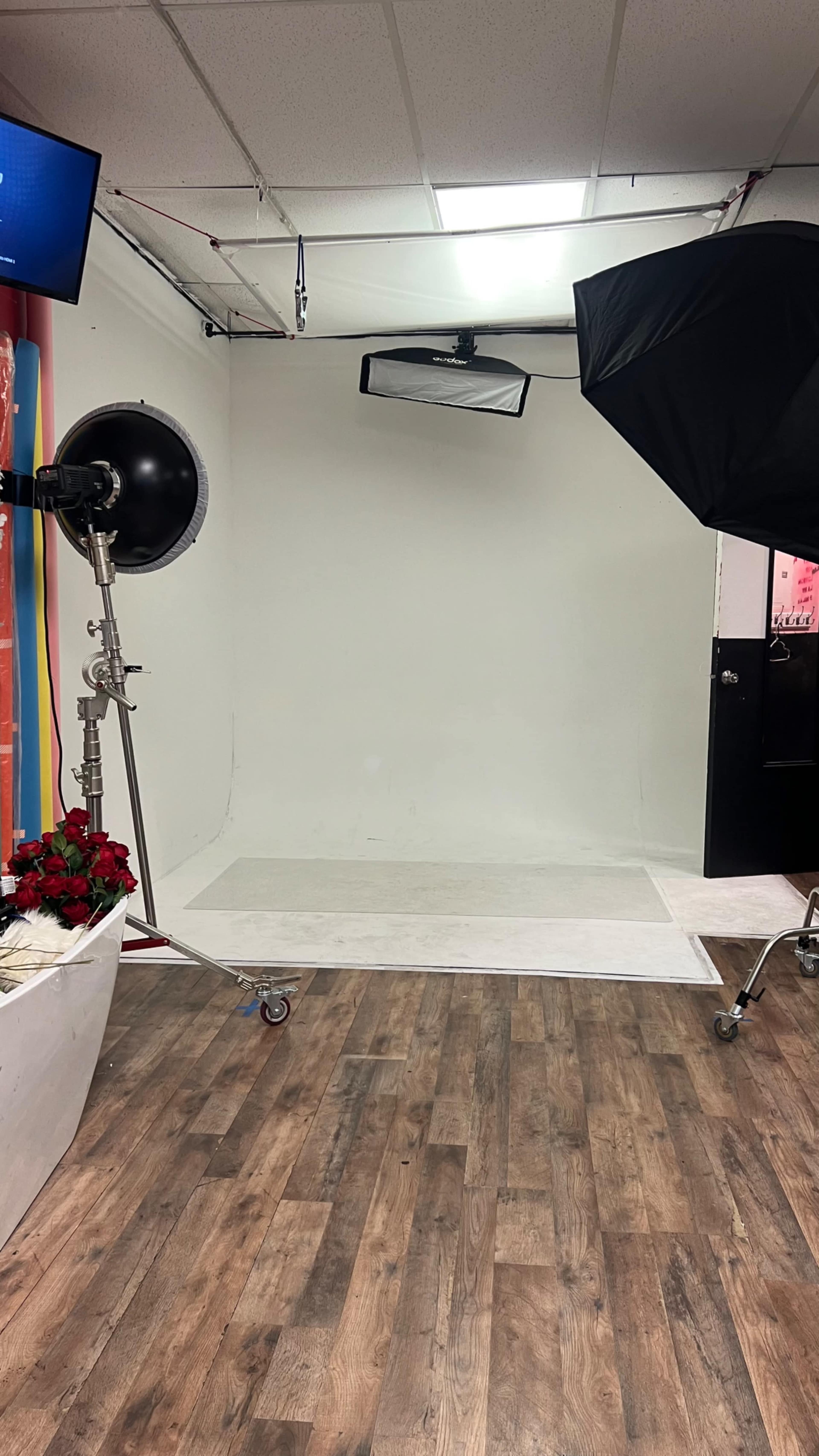A photography studio features a blank white backdrop, lighting equipment, and a cart with roses on the floor.