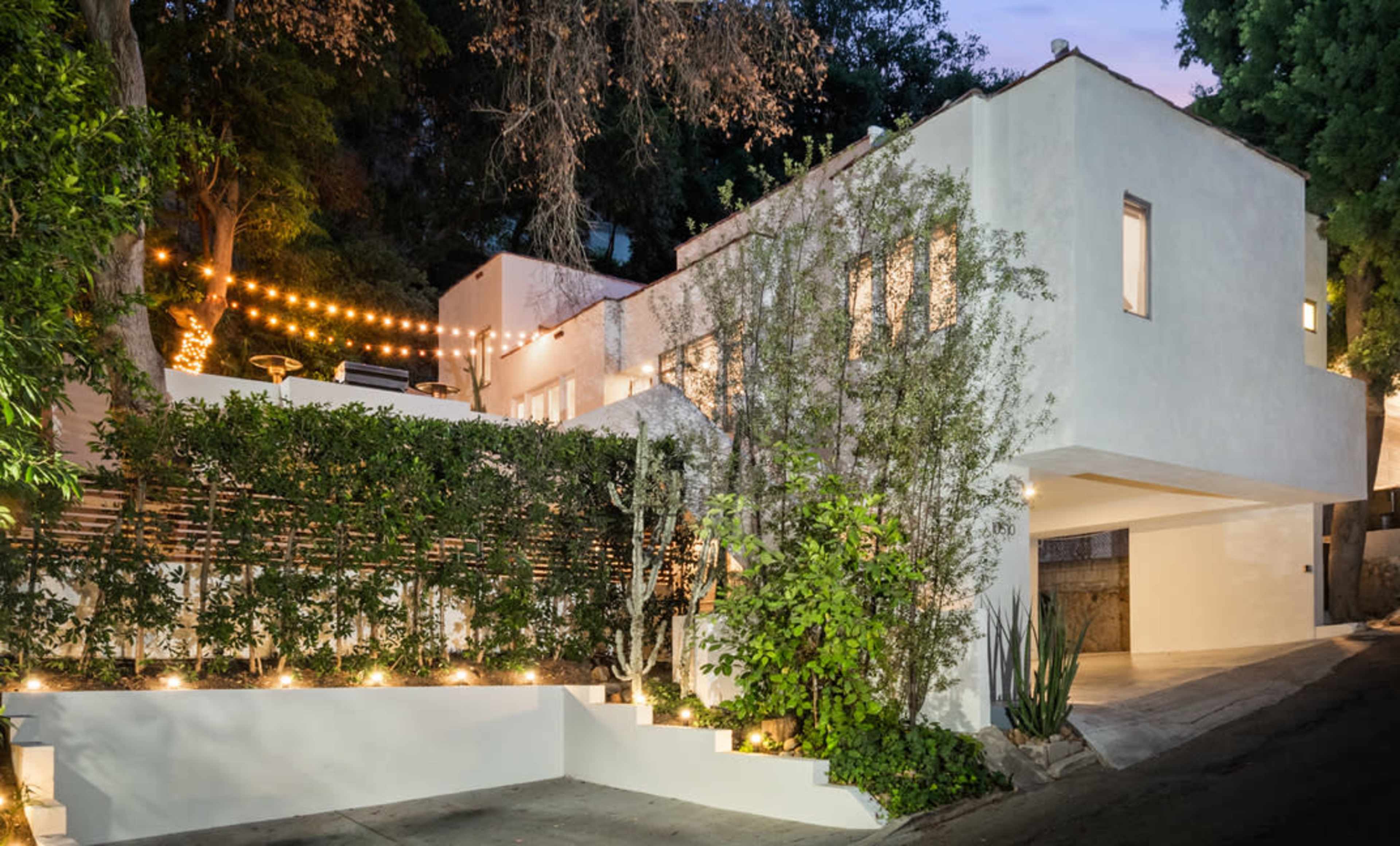 A modern white house on a hillside is bordered by greenery and strung with lights, creating a welcoming atmosphere in the evening.