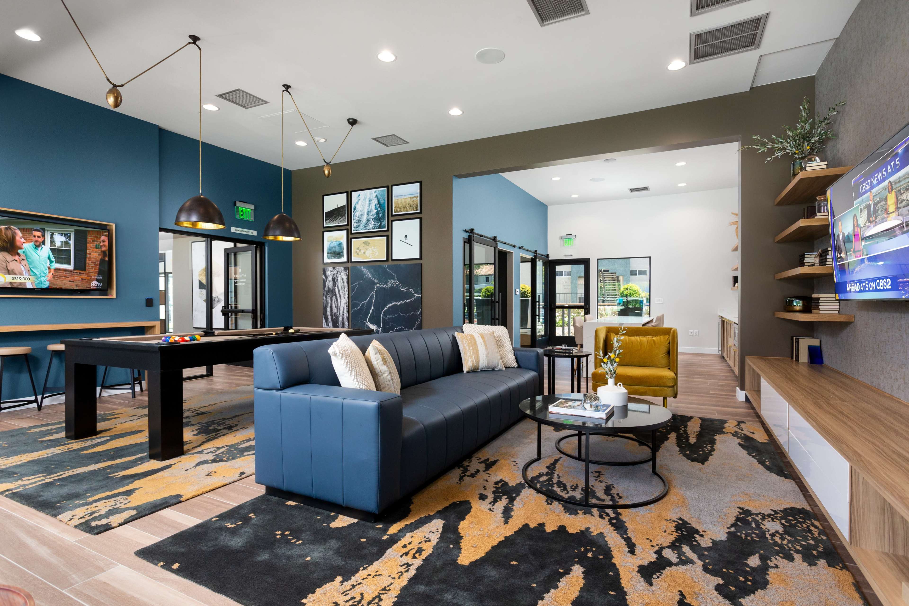 A modern lounge area with a blue sofa, a pool table, and a large television mounted on the wall, surrounded by decorative artwork and plants.