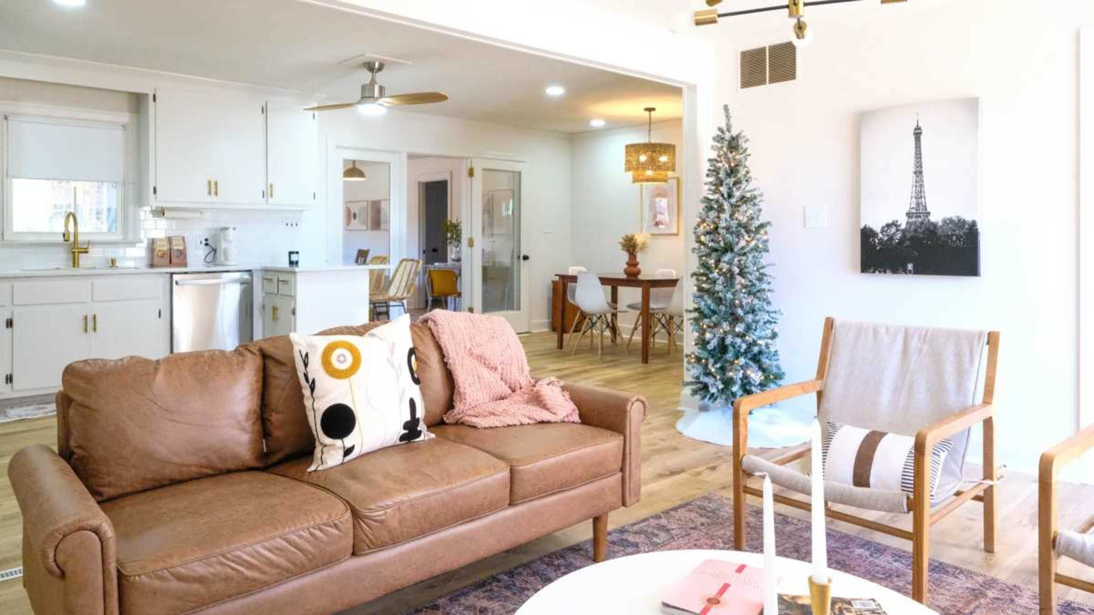 A cozy living room features a brown leather sofa with decorative pillows, a Christmas tree in the corner, and a dining area visible in the background.