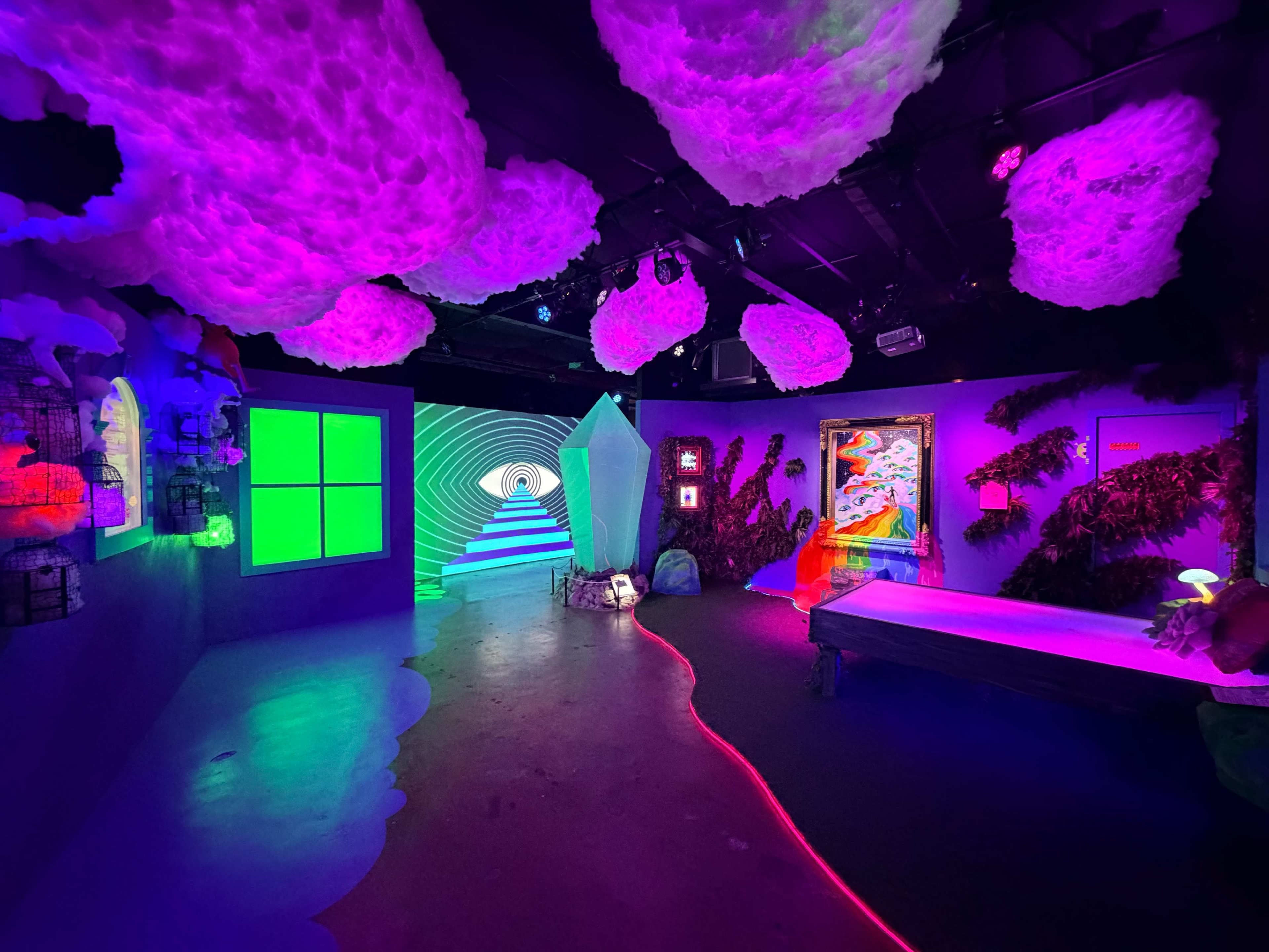 The image shows a vibrant and colorful room with neon lighting, featuring a large geometric crystal, illuminated cloud-like decorations overhead, and various artworks on the walls.