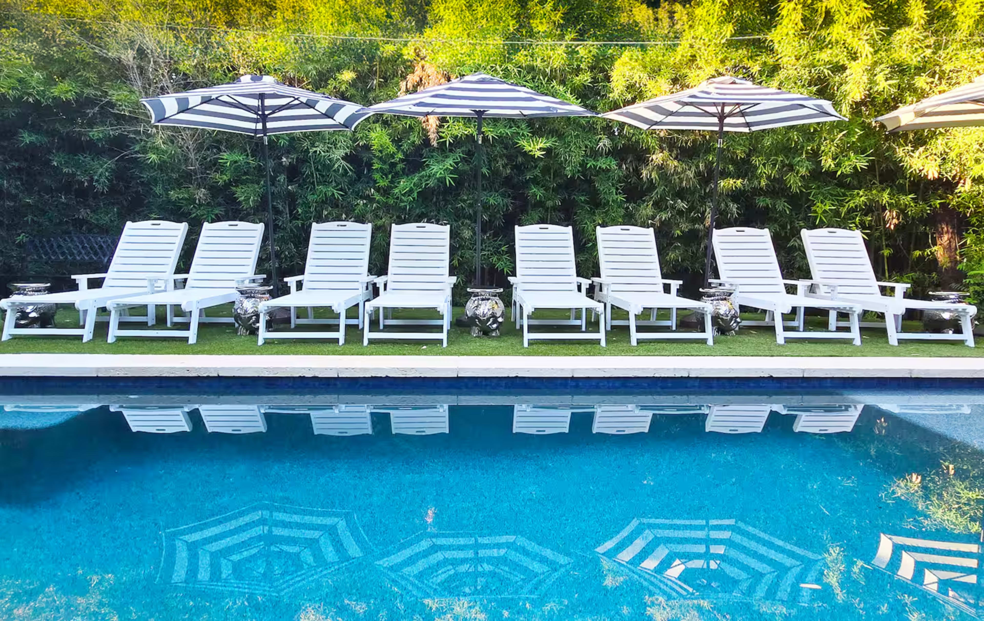 A row of white lounge chairs and umbrellas is positioned beside a swimming pool, with their reflections visible on the water's surface.