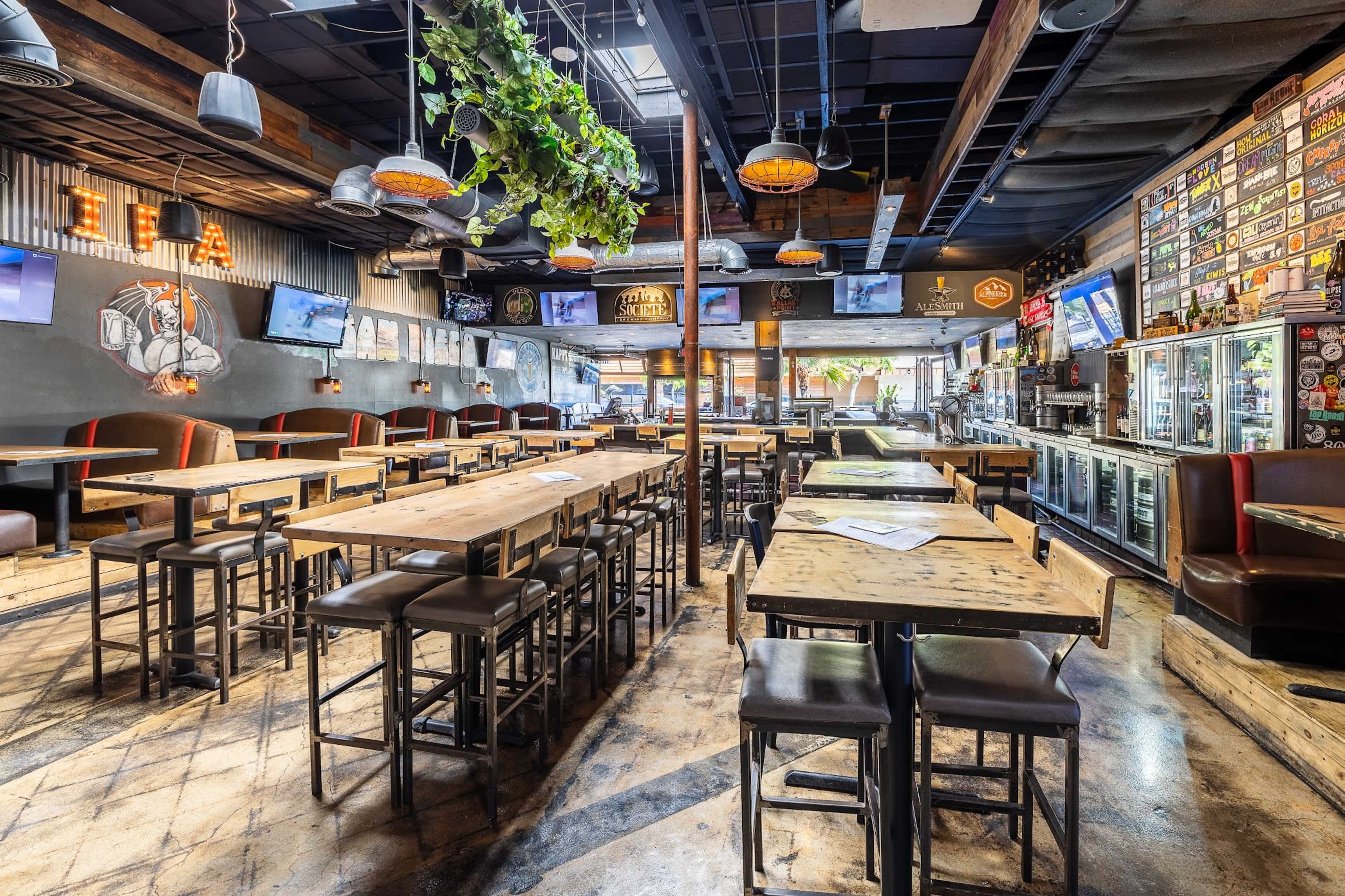 The image shows an interior view of a restaurant or bar featuring multiple wooden tables, high stools, and a long counter with a drink cooler, surrounded by walls adorned with decorations and monitors.