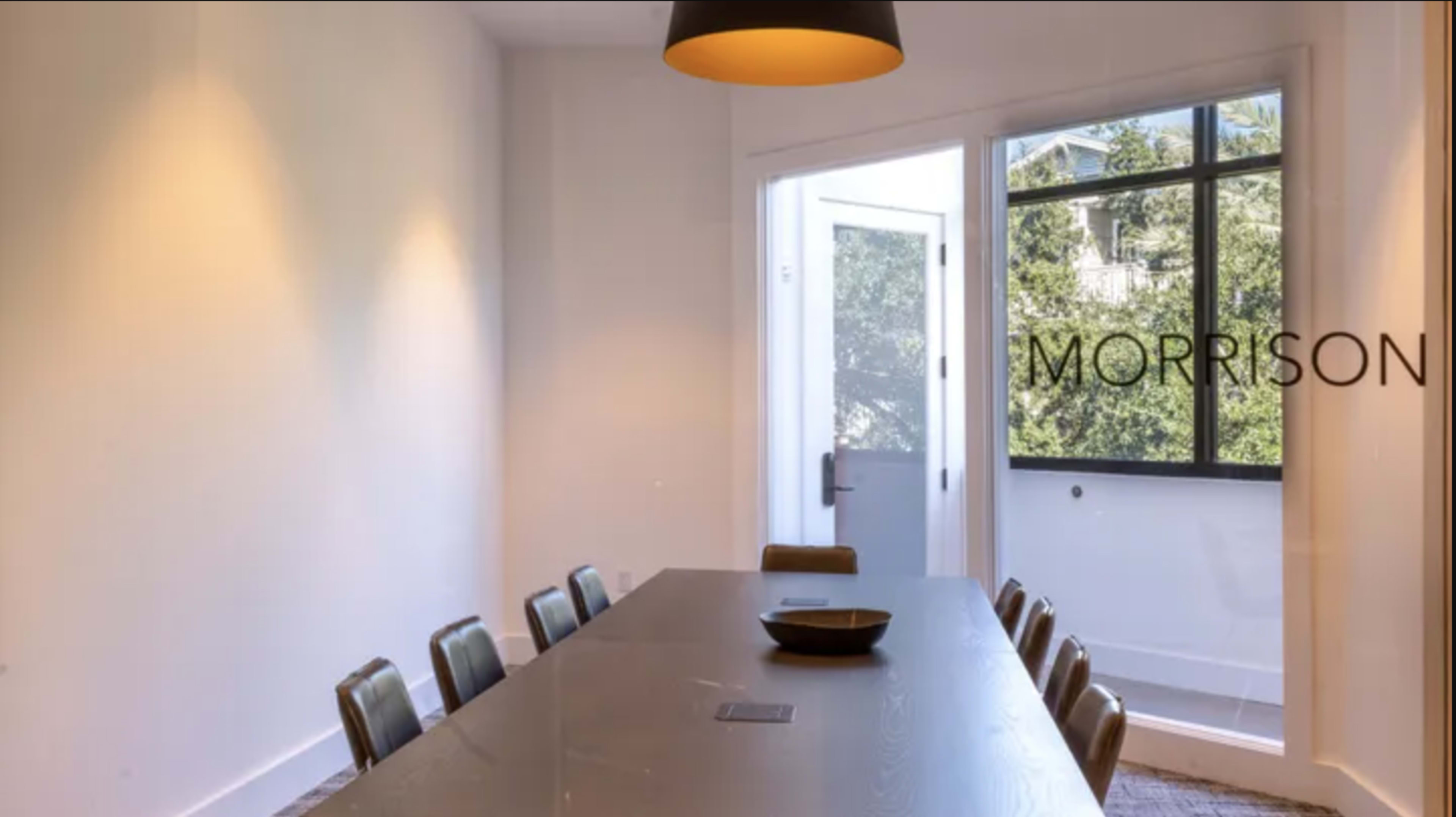 A long table with chairs is set in a well-lit room featuring a large window that opens to an outdoor view.