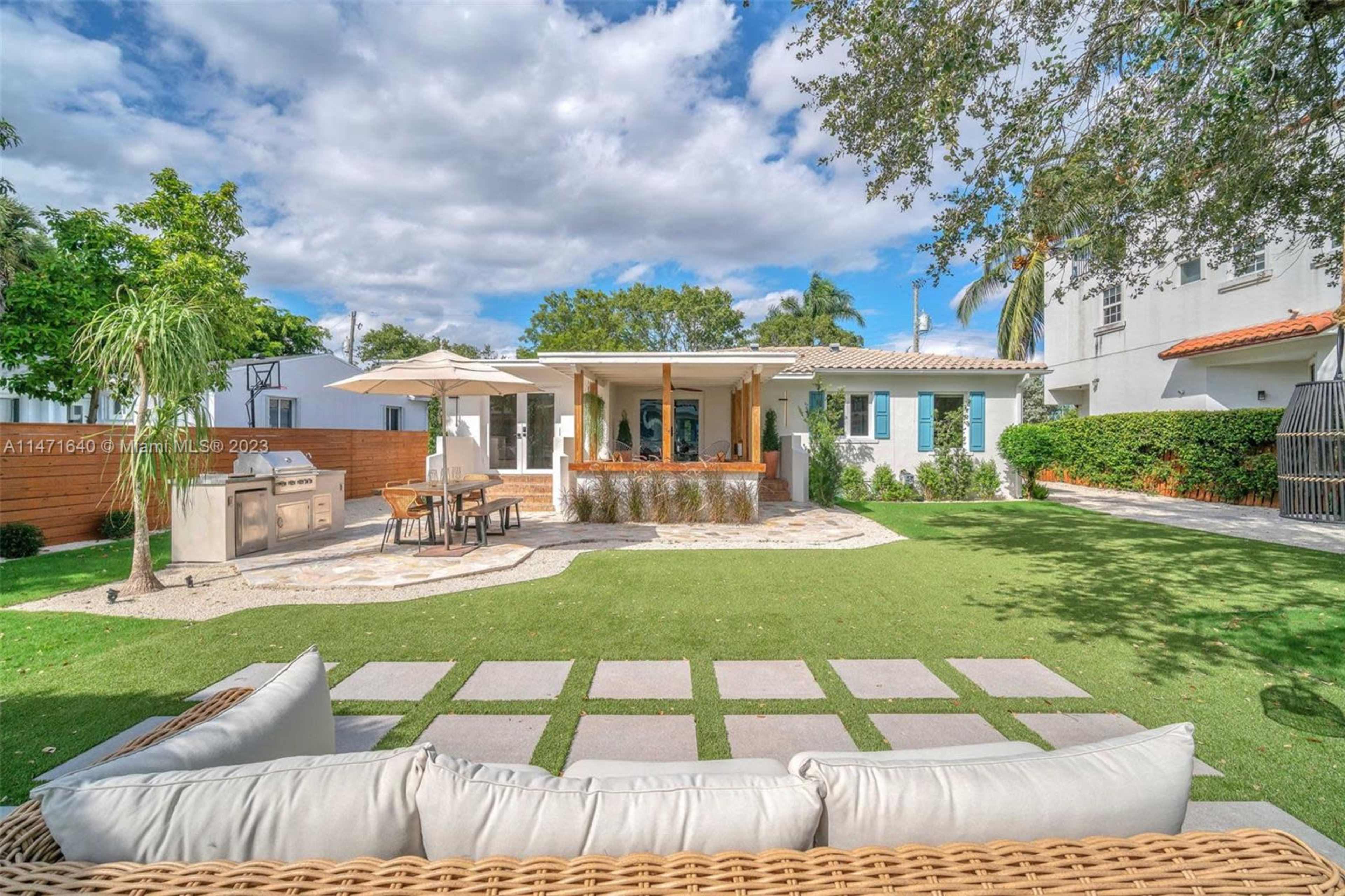 The image shows a residential backyard featuring a grassy area with stone pavers, a covered patio with outdoor seating, and a landscaping of trees and shrubs.