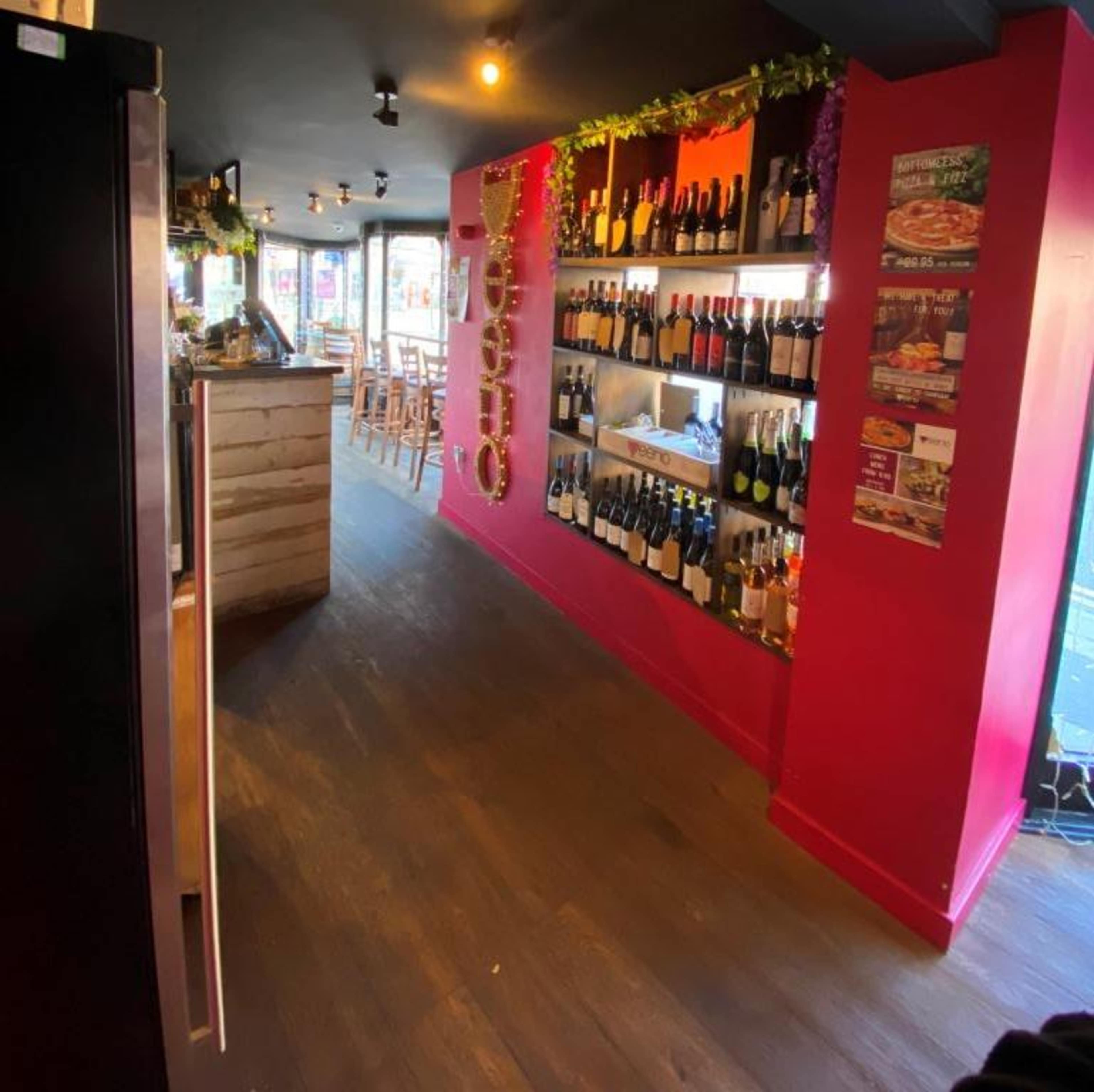 The image shows a bar area with a display of wine bottles on shelves against a bright pink wall, and seating along one side.