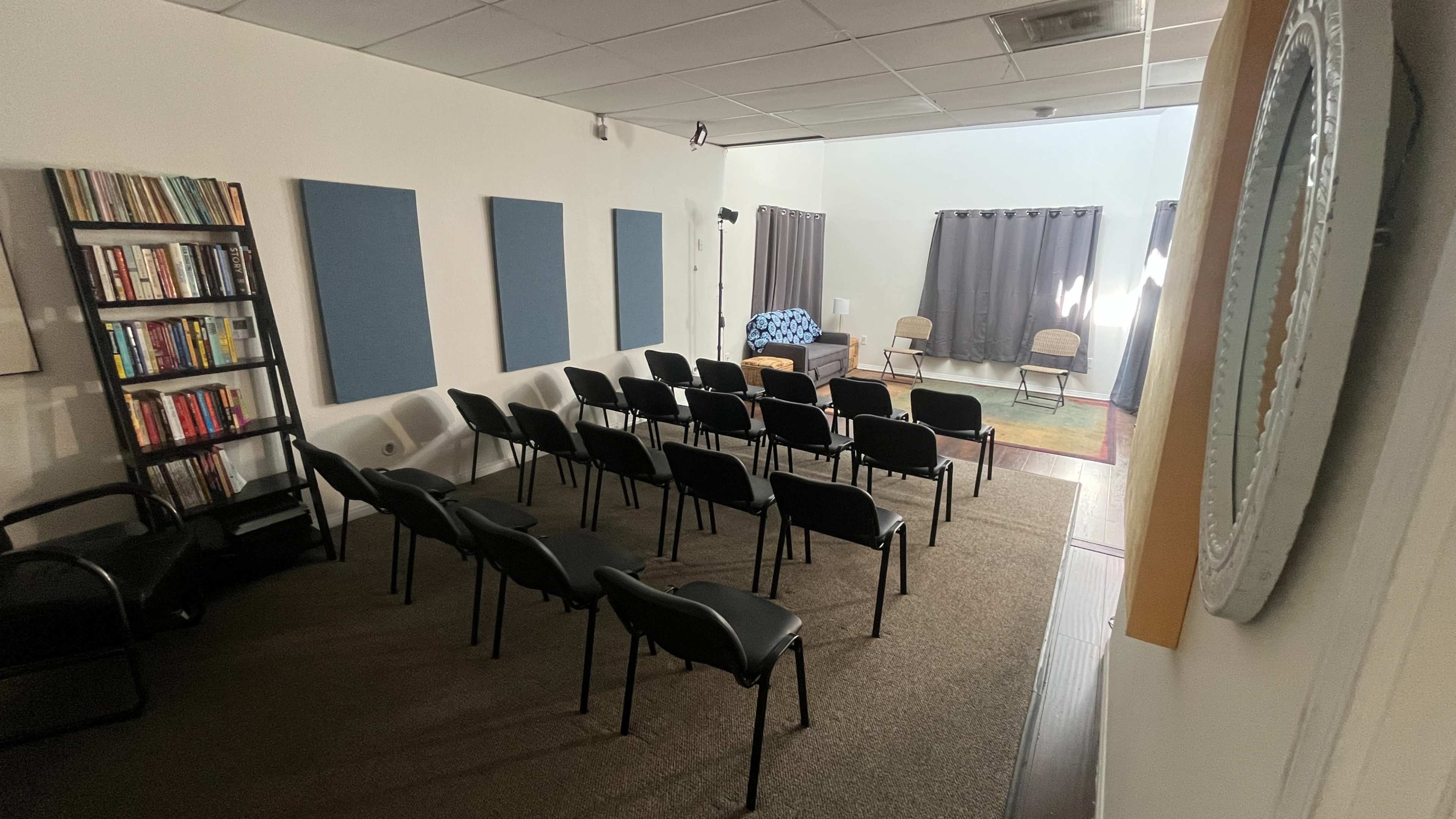 The image shows a small meeting room with rows of black chairs facing a stage area that has curtains, a rug, and a few chairs.