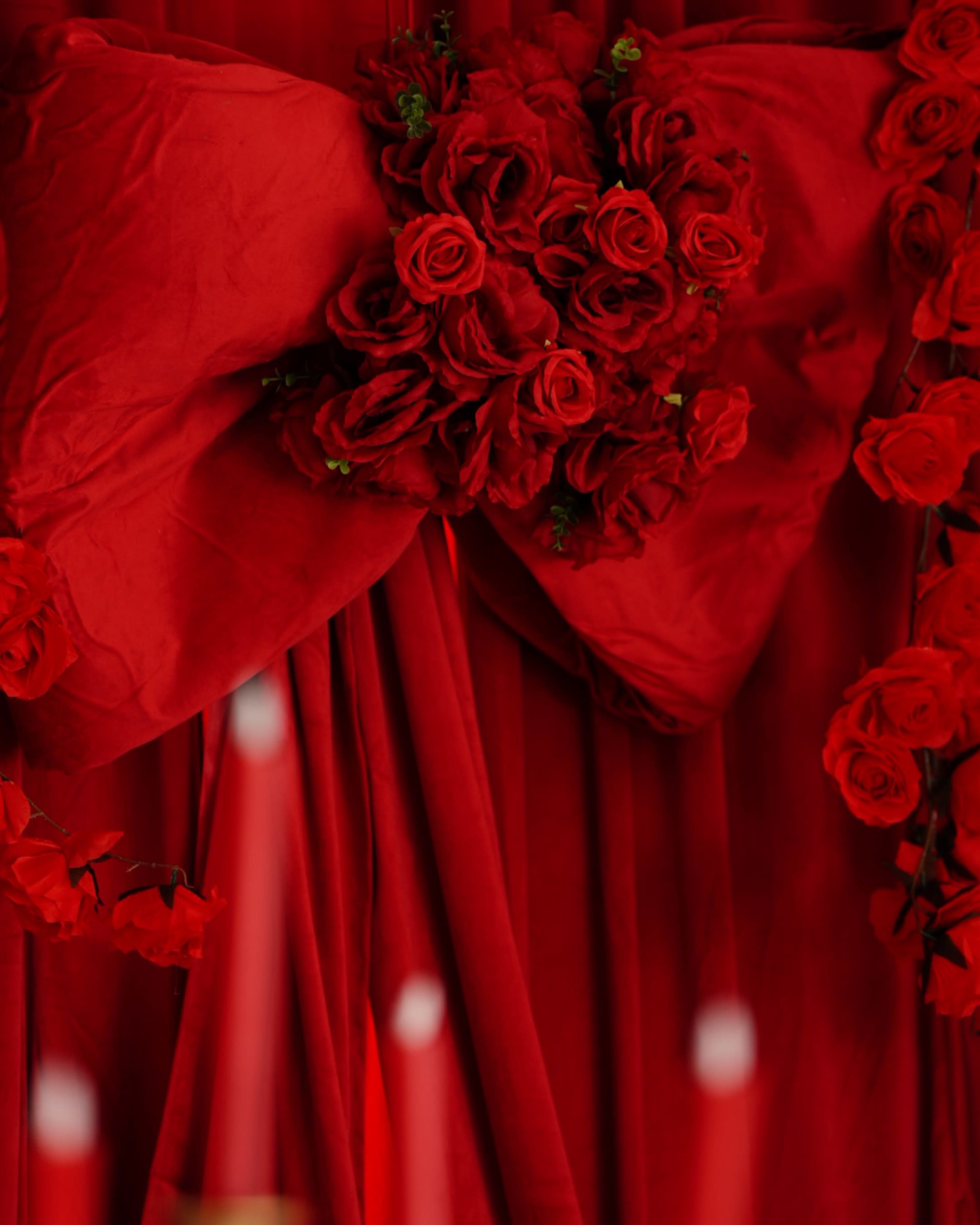 A large red bow adorned with artificial roses is displayed against a backdrop of red fabric.