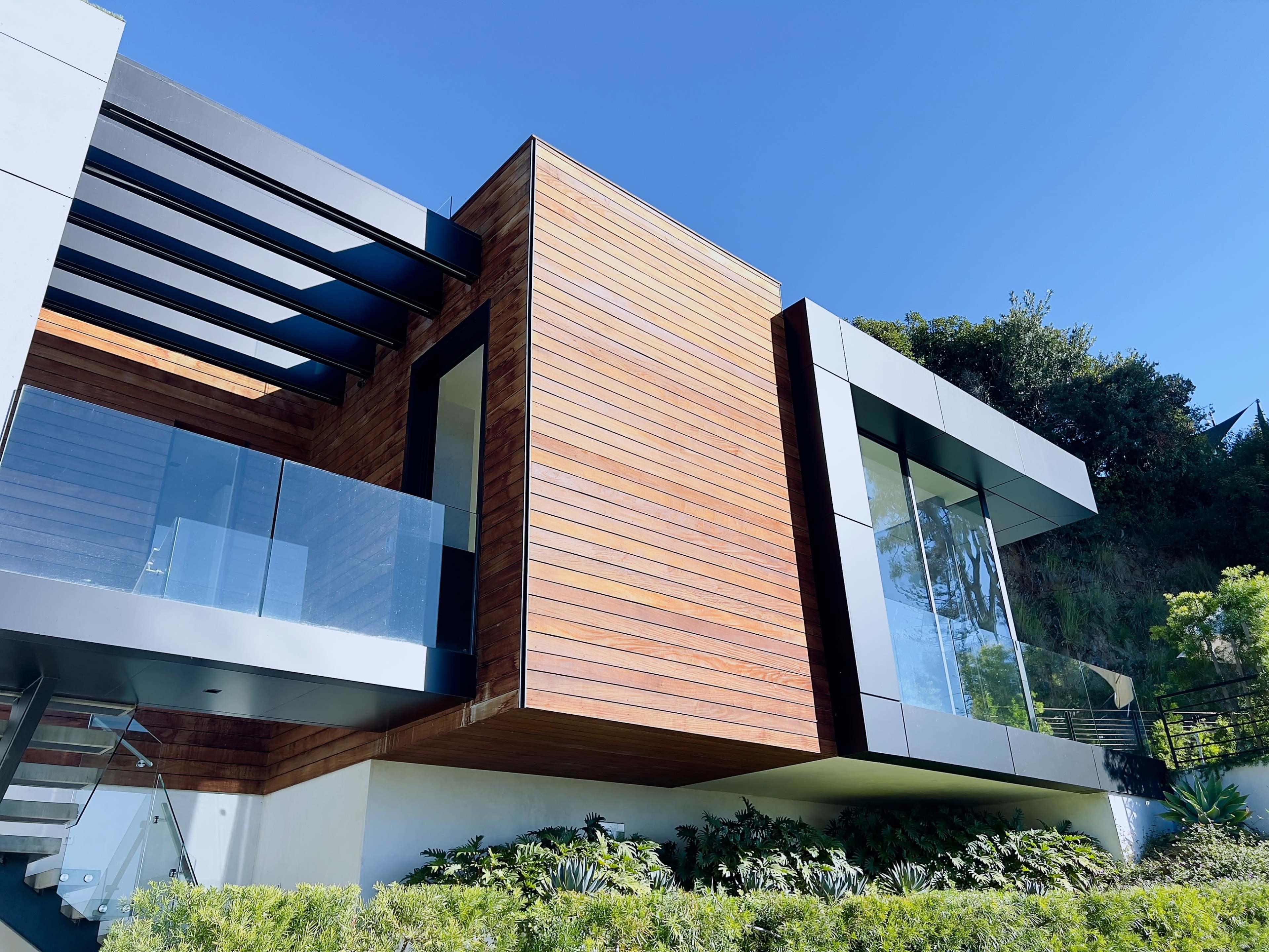 The image features a modern house with a wooden exterior, large glass windows, and a sleek design set against a clear blue sky.