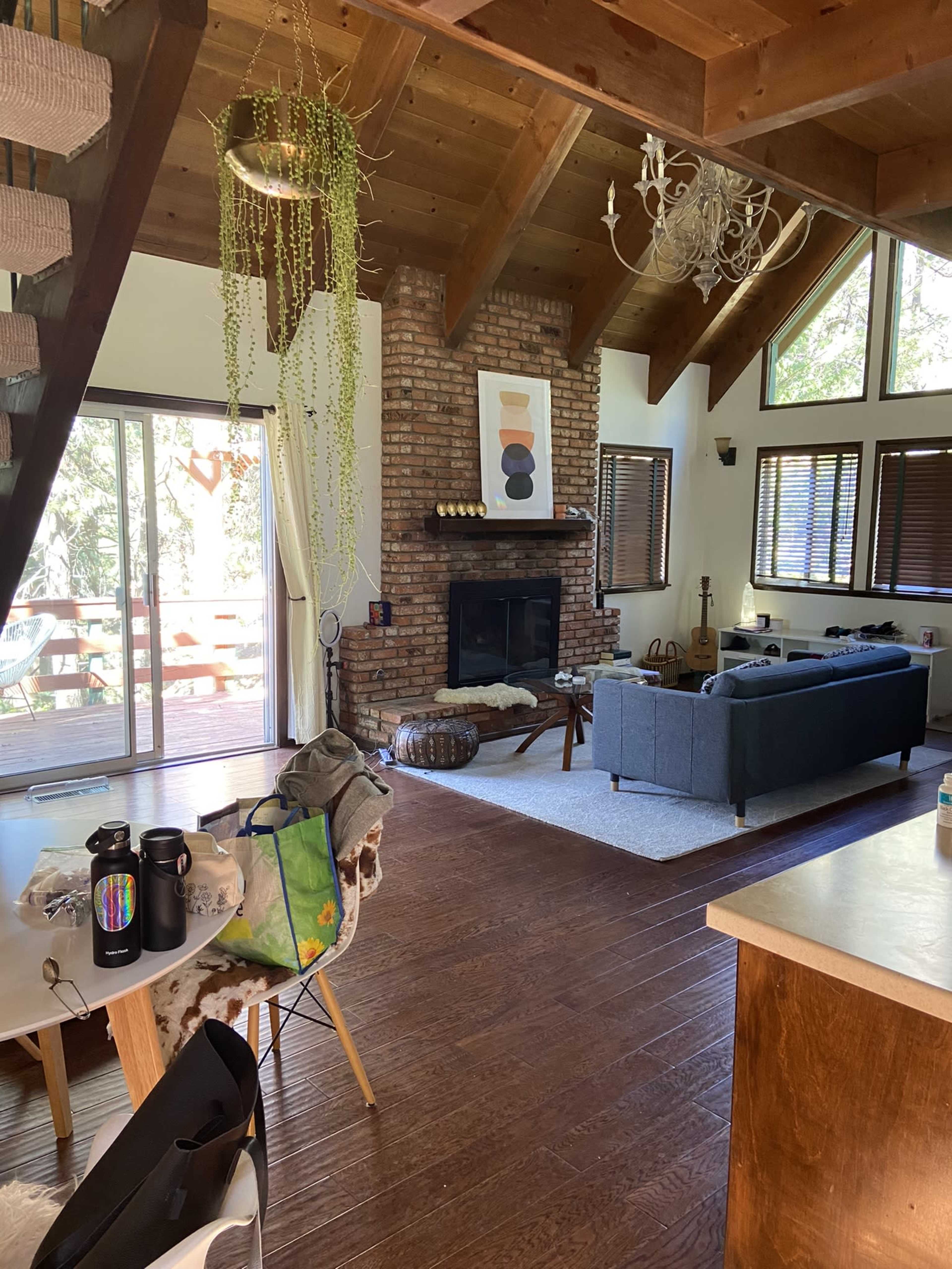 A living room with a brick fireplace, wooden beams, and large windows overlooking a deck, furnished with a sofa and various personal items scattered around.