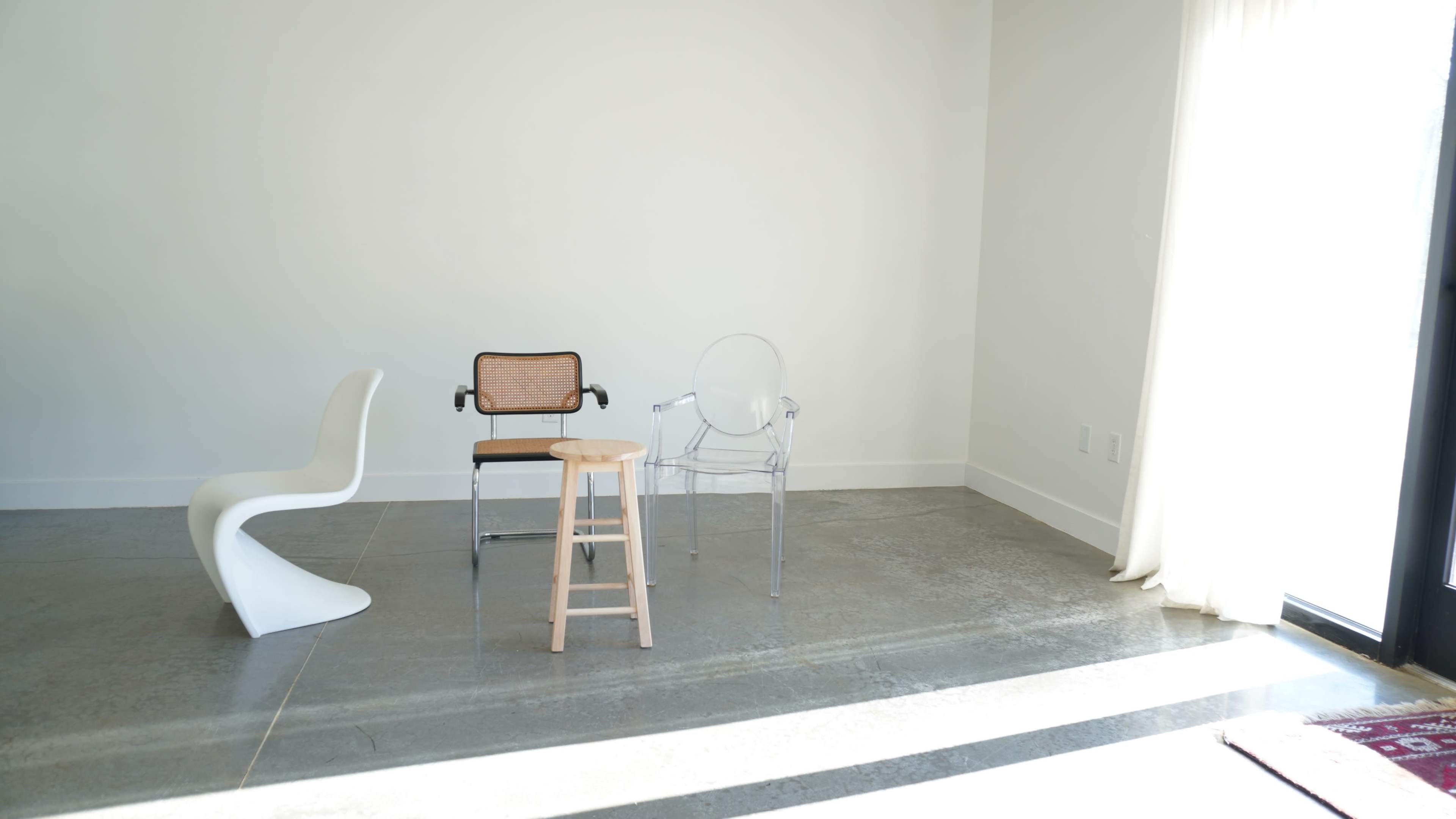 The image shows three different chairs positioned on a light gray floor in a minimally furnished room with white walls and natural light coming through a window.