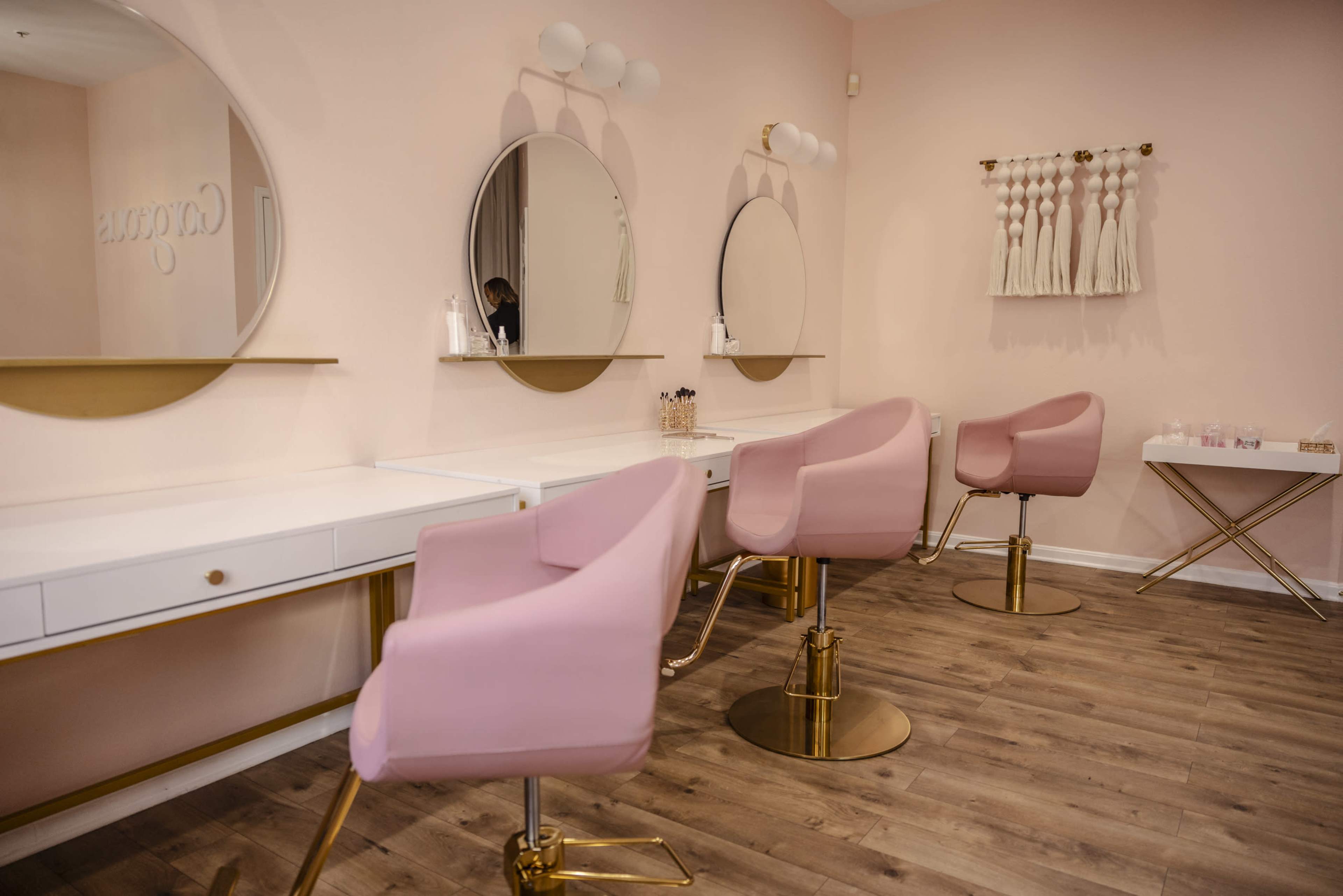 A modern beauty salon with pink walls, two circular mirrors above white vanities, and pink chairs with gold accents.