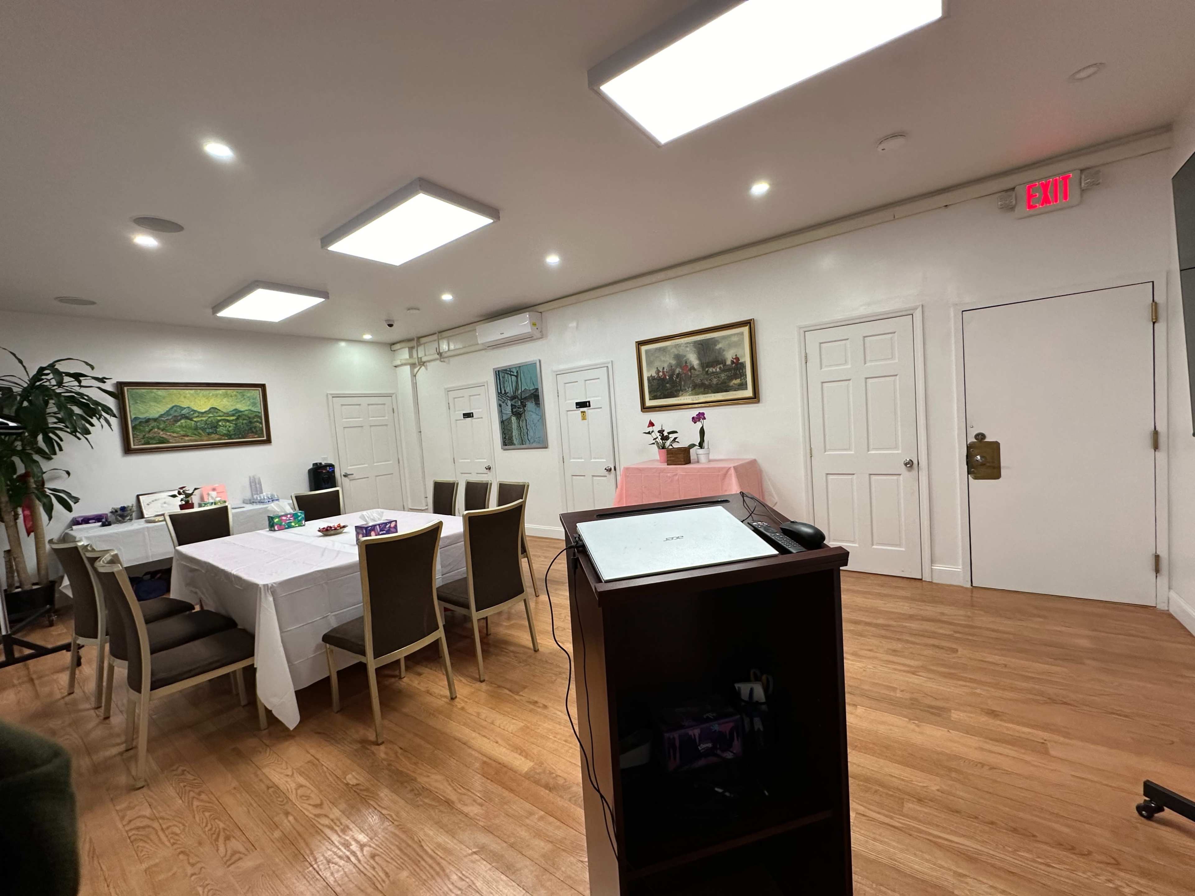 The image shows a spacious room setup for a gathering, featuring several tables covered with white tablecloths, chairs arranged around them, and decorative wall art.