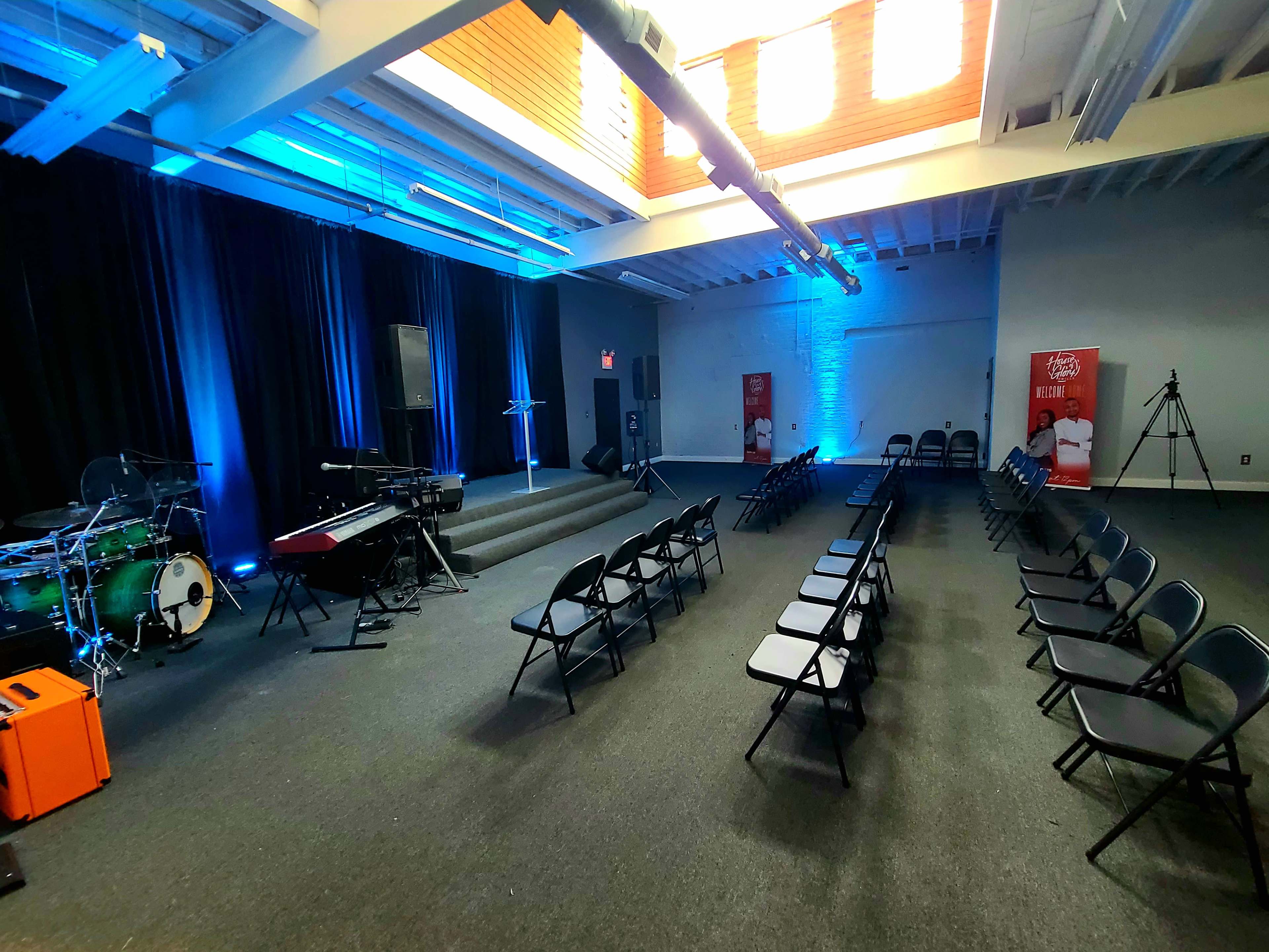 A room set up for an event, featuring rows of black chairs facing a stage with musical instruments and blue lighting.