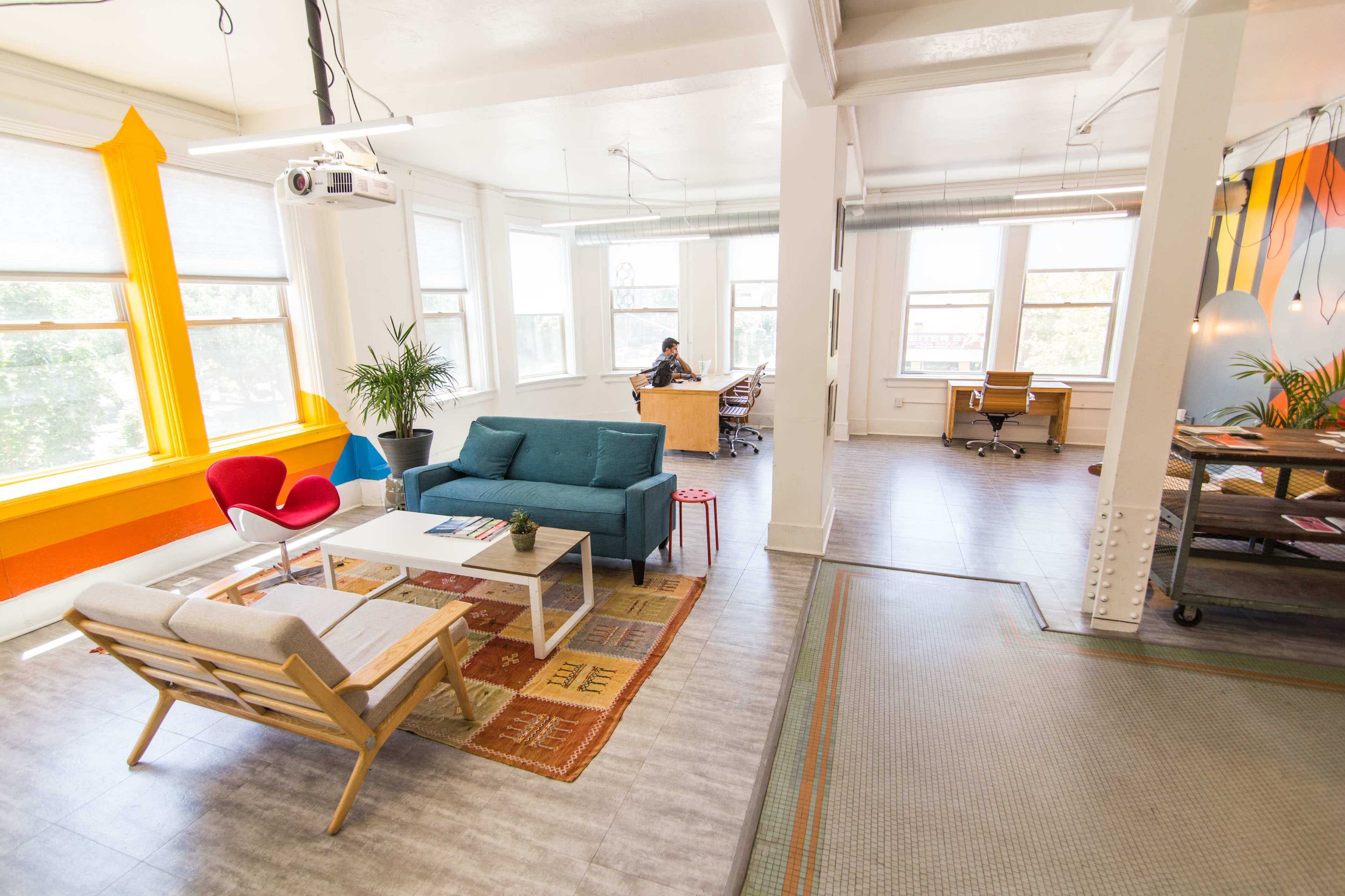 A bright and modern co-working space with a seating area featuring a blue sofa, a red chair, and a coffee table, and an adjacent area with desks and a projector.