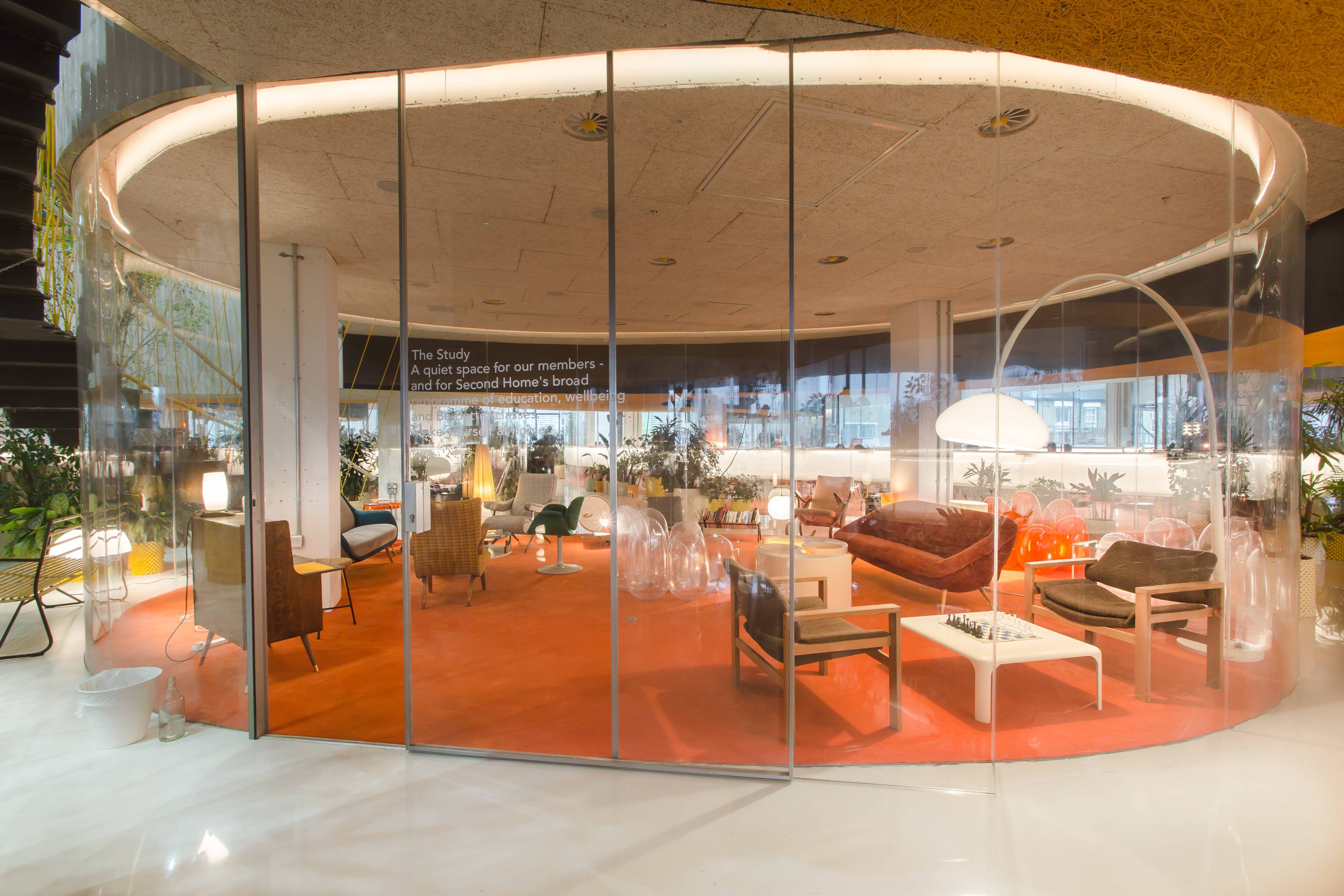 A modern, circular lounge area with glass walls, featuring various seating arrangements and plants.