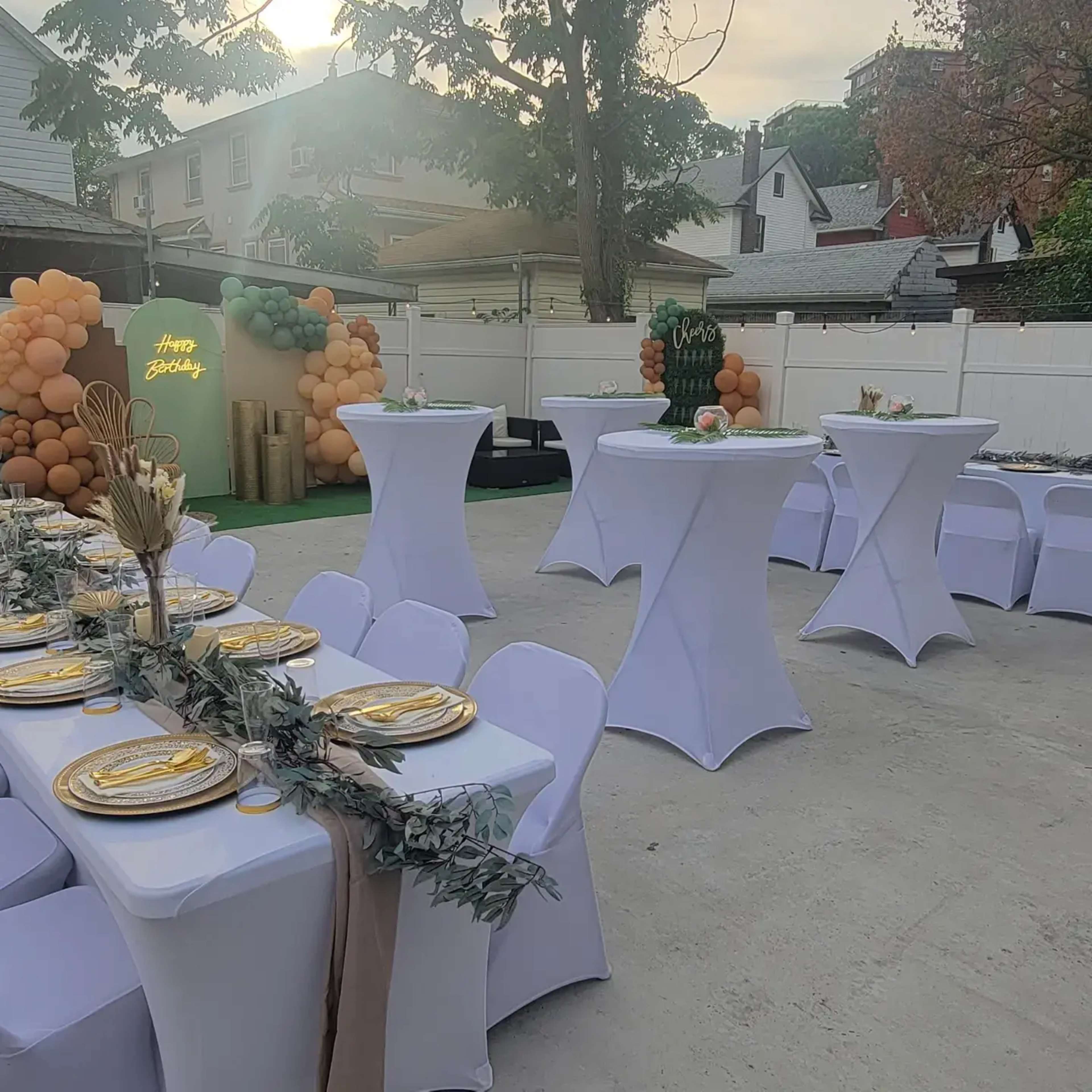 The image shows a decorated outdoor party area featuring tables with white tablecloths, golden plates, and balloon decorations in varying colors.