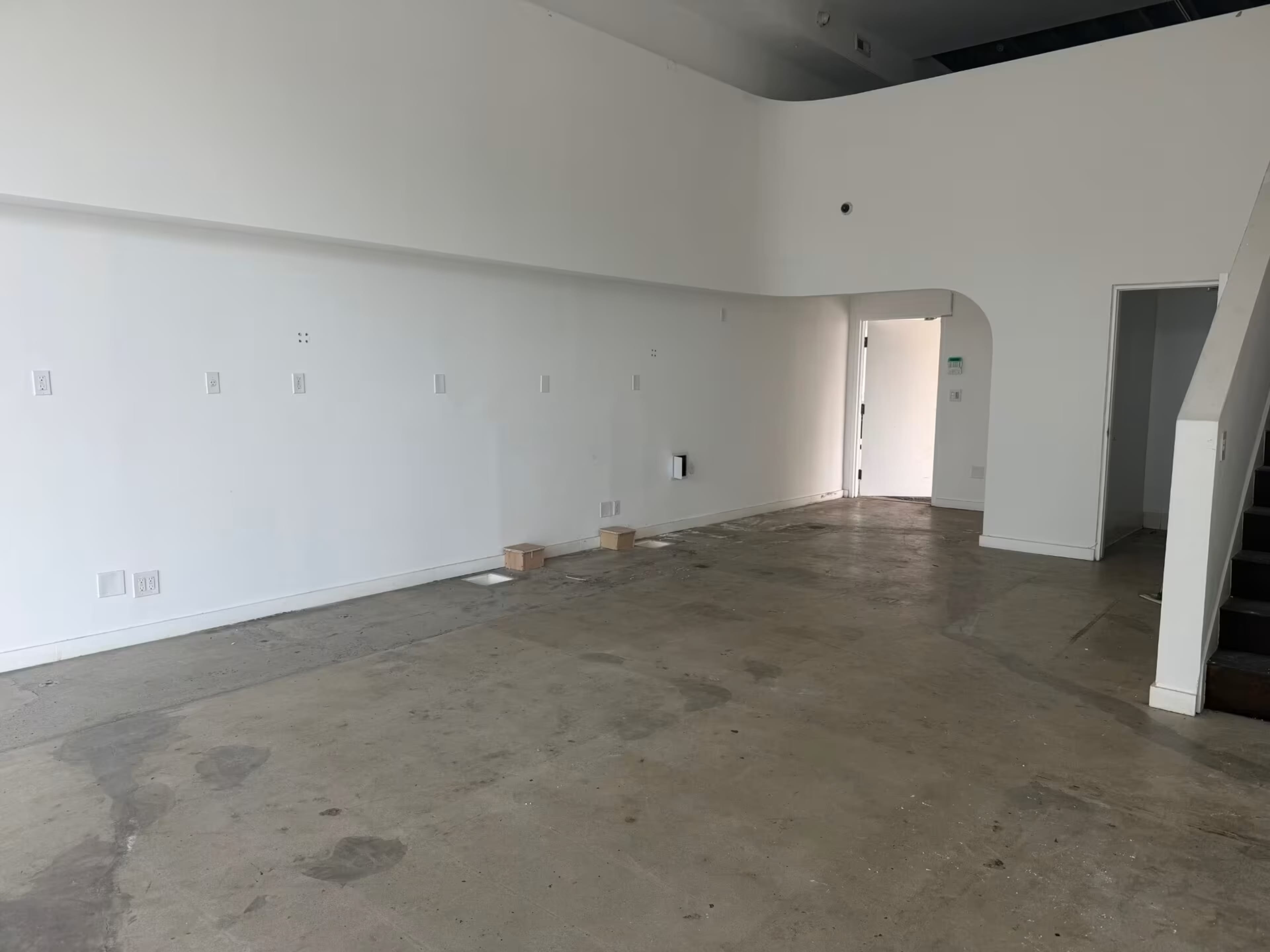 The image shows a large, empty room with white walls, concrete flooring, and a staircase on the right side.