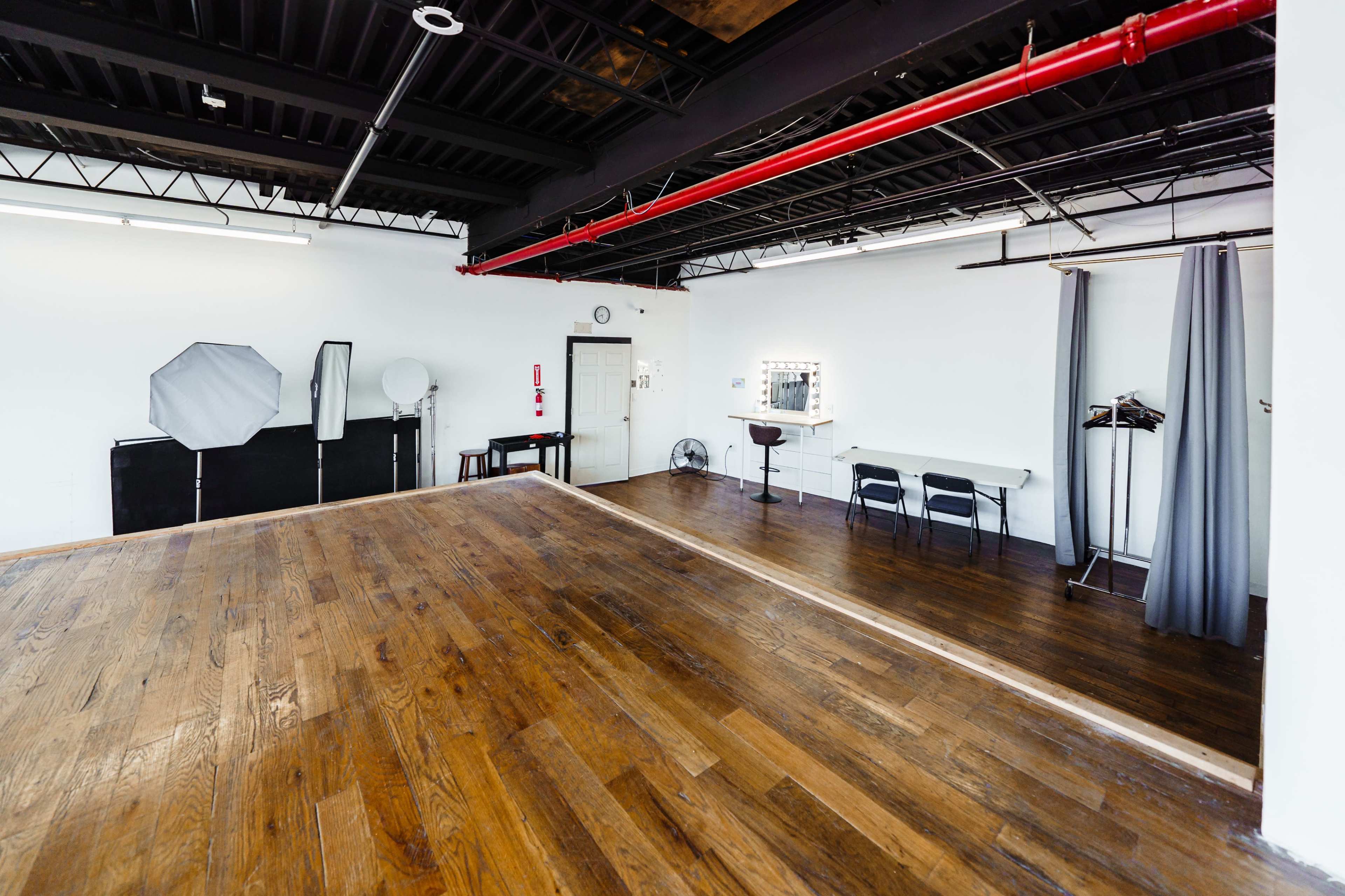 A spacious studio with wooden flooring, equipped with a mirror, chairs, and various photography equipment against the walls.