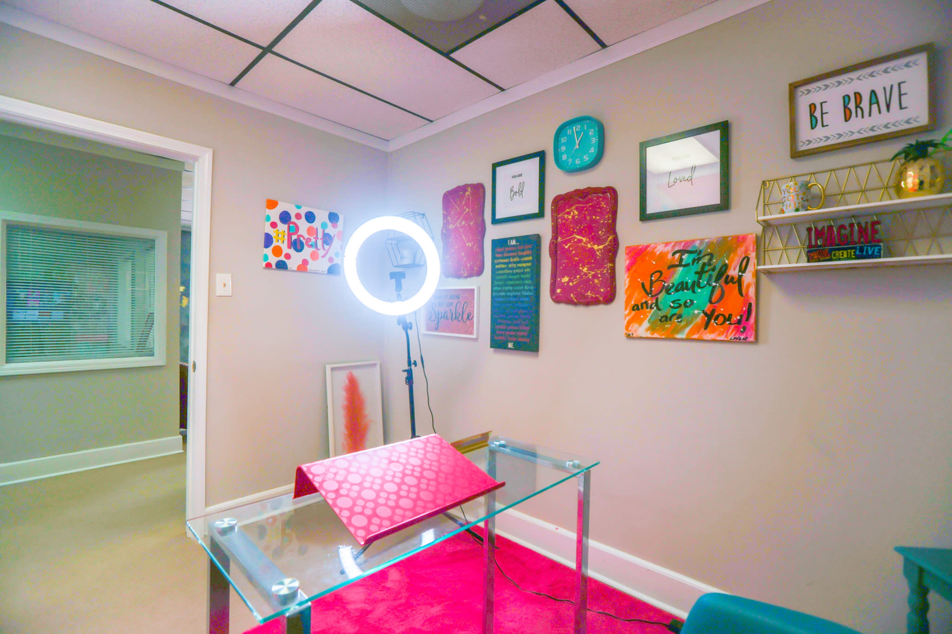 A small office space featuring a ring light on a tripod, a glass table with a patterned cloth, and various colorful artworks displayed on the walls.