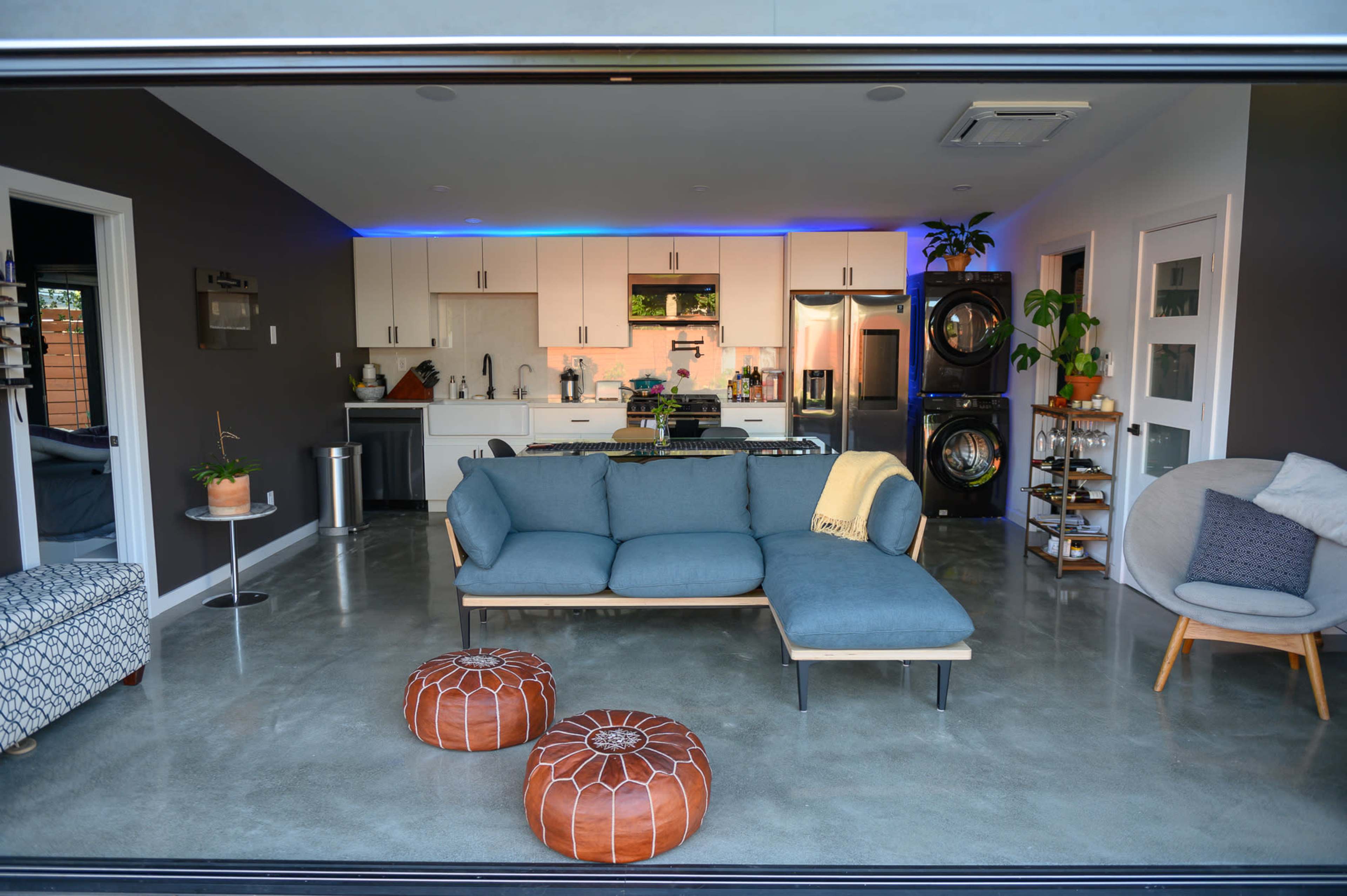 A modern living room features a blue sectional sofa, two round poufs, and a kitchen visible in the background, all set against a minimalist concrete floor.