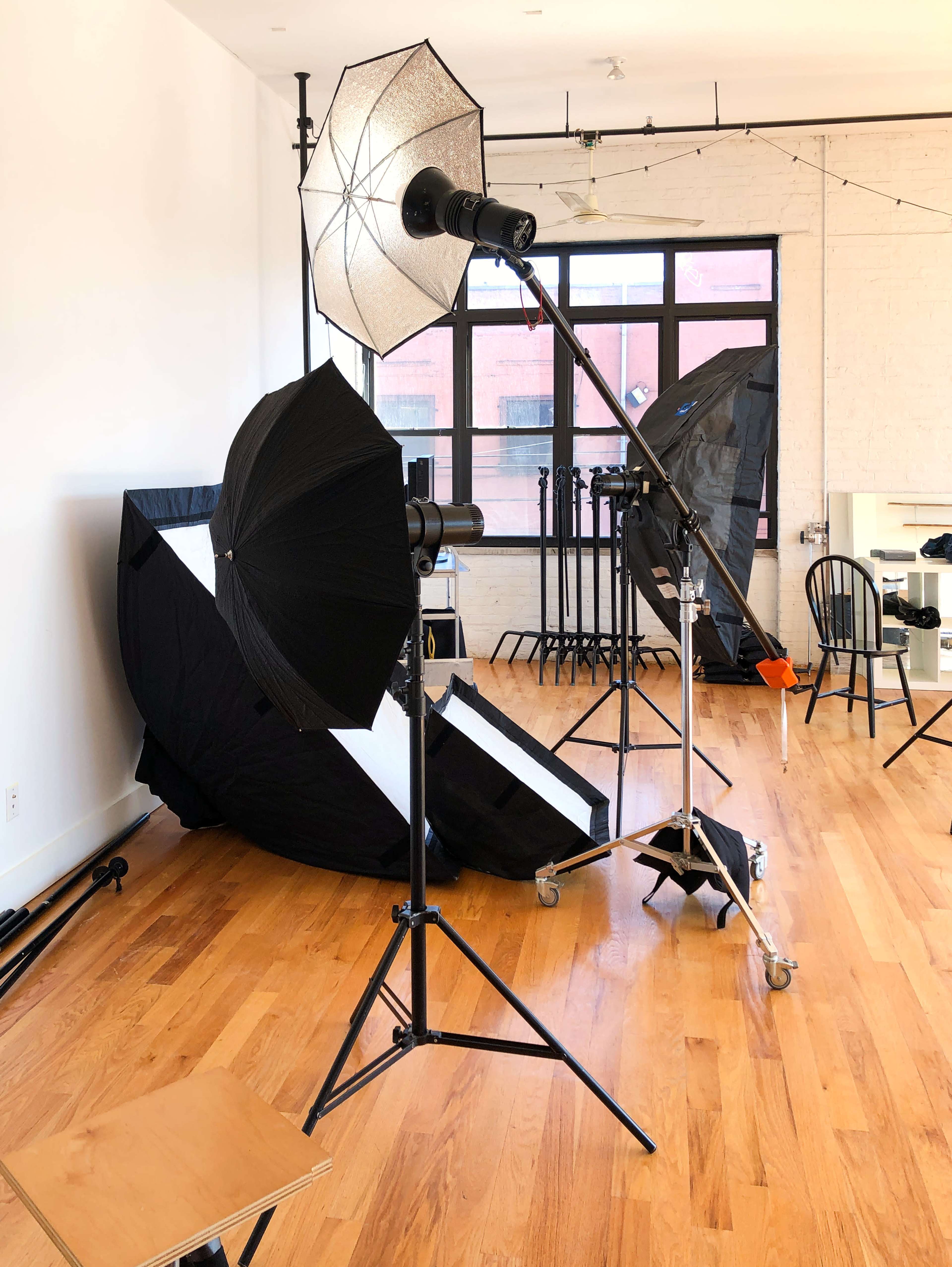 A photography studio with lighting equipment set up, including umbrellas and softboxes, against a backdrop of wooden flooring and large windows.