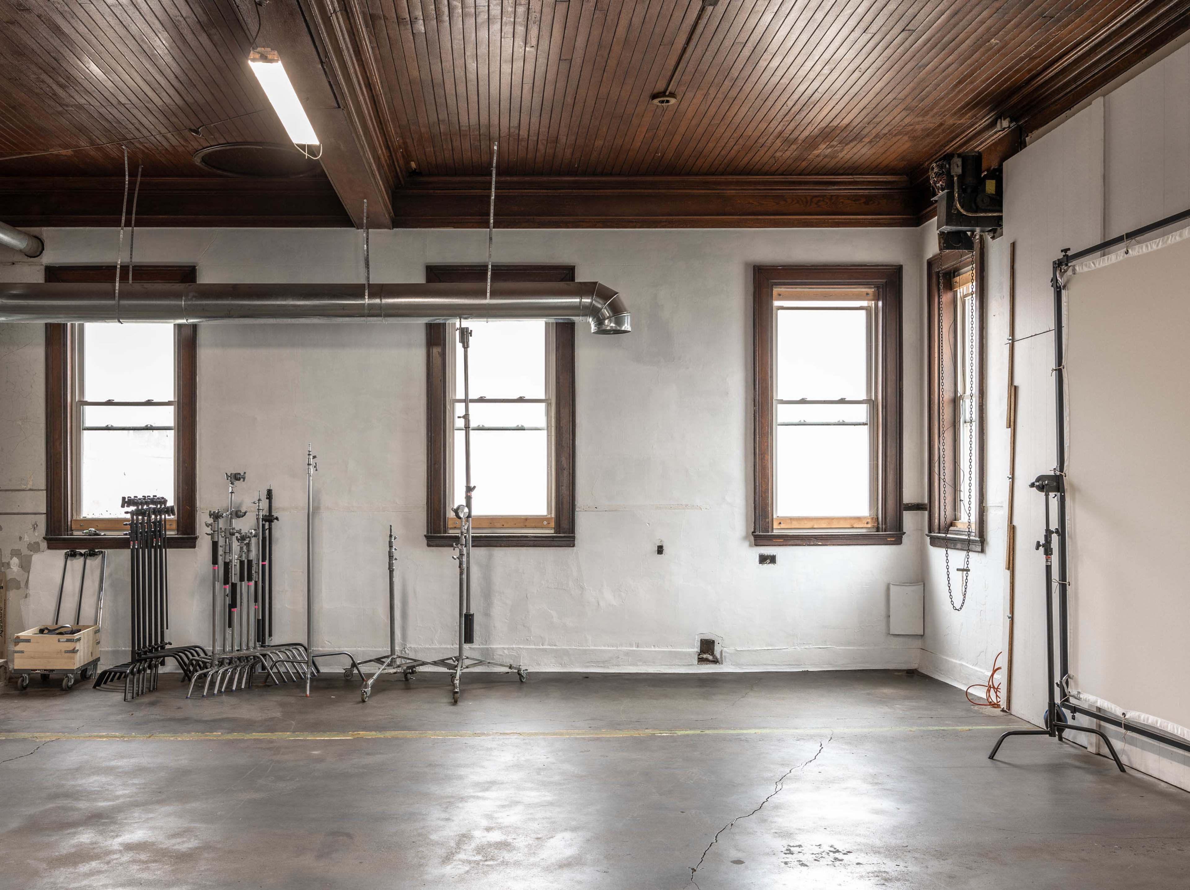 The image shows a spacious, empty room with wooden ceiling beams, two windows, and several metal stands positioned against the walls.