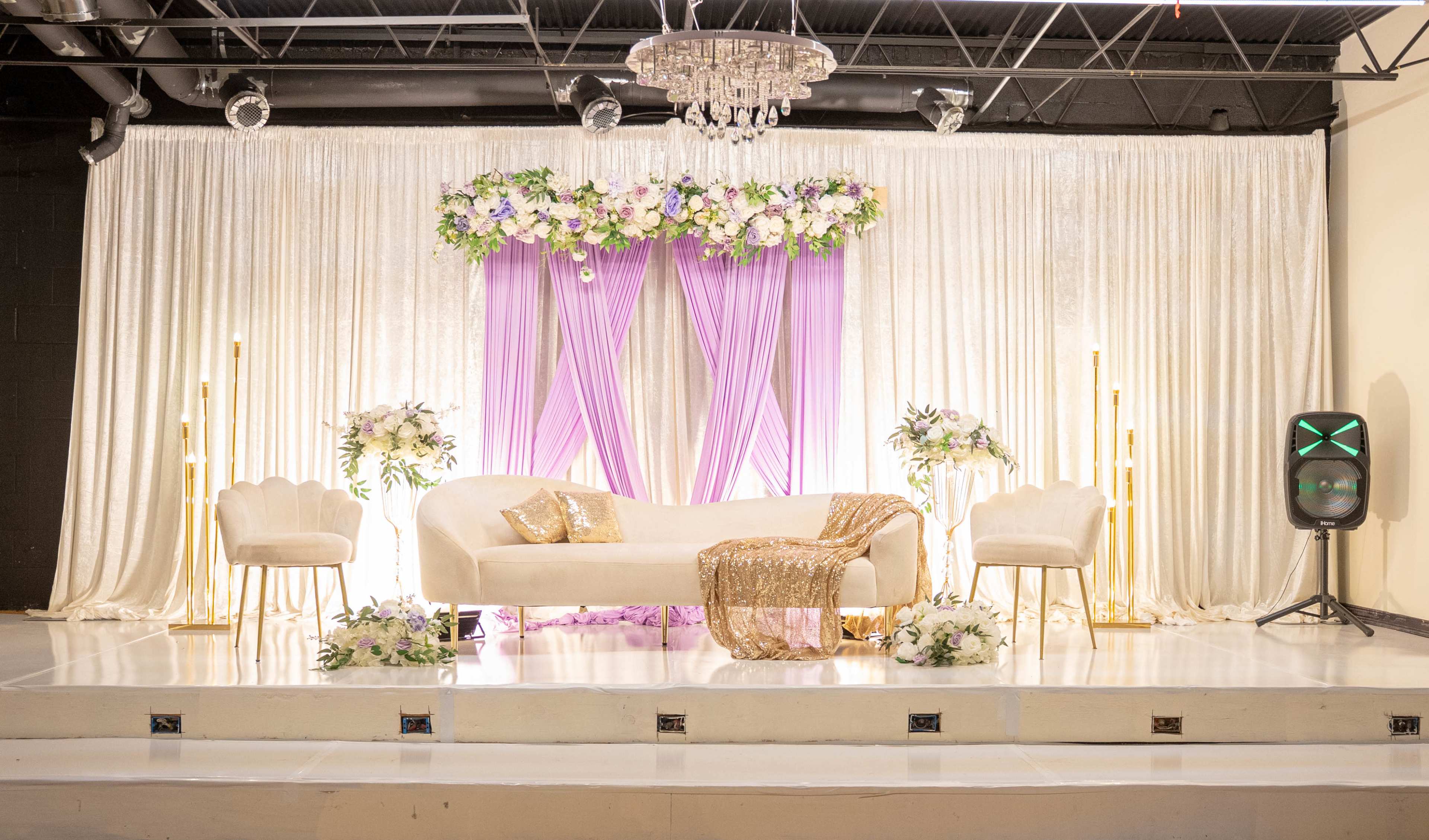 The image shows a decorated stage featuring a white sofa surrounded by floral arrangements, draped curtains in shades of purple and gold, and accent chairs with a chandelier overhead.