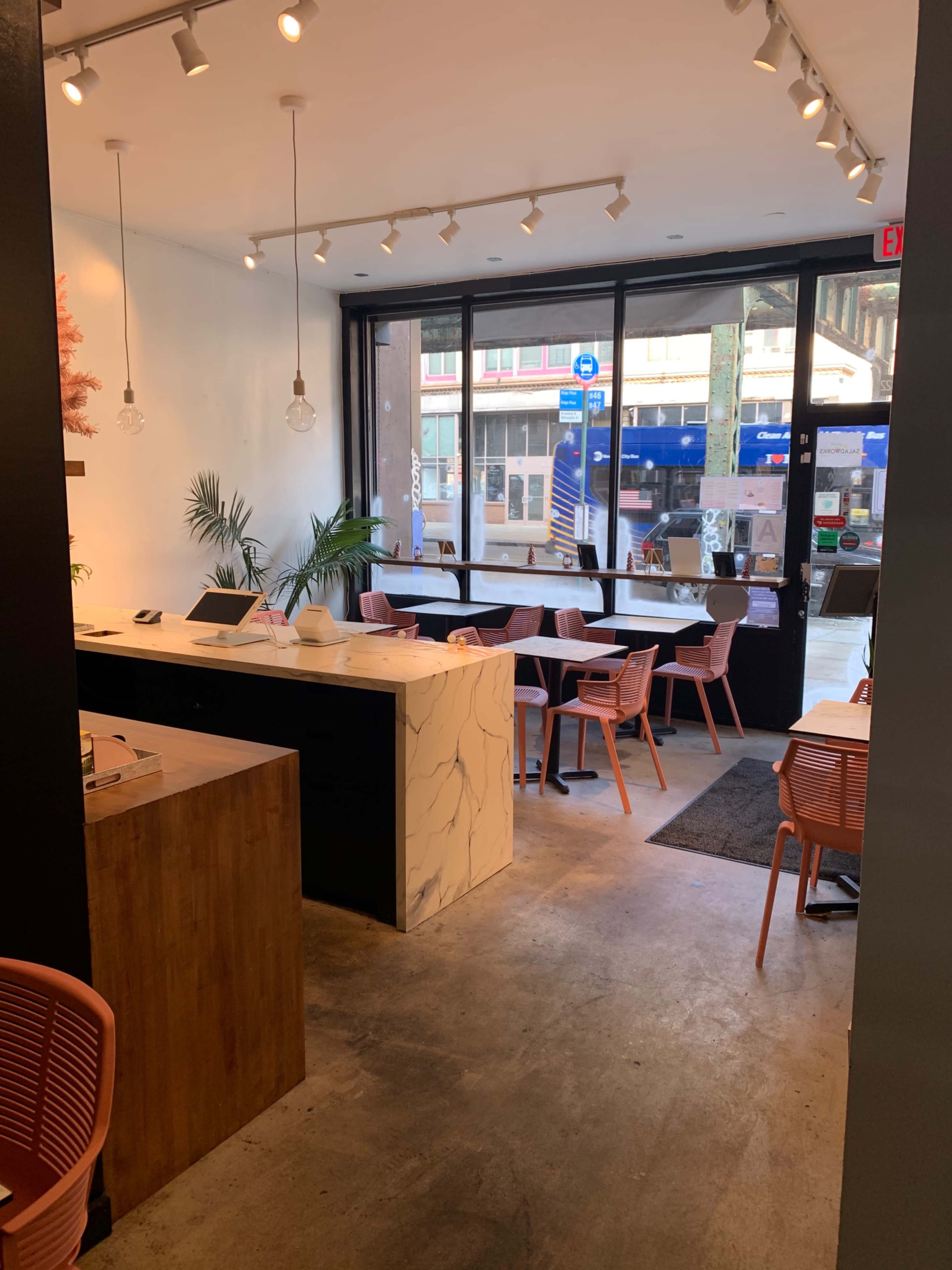 The interior of a cafe features a minimalist design with a marble counter and pink chairs, illuminated by natural light from large windows.