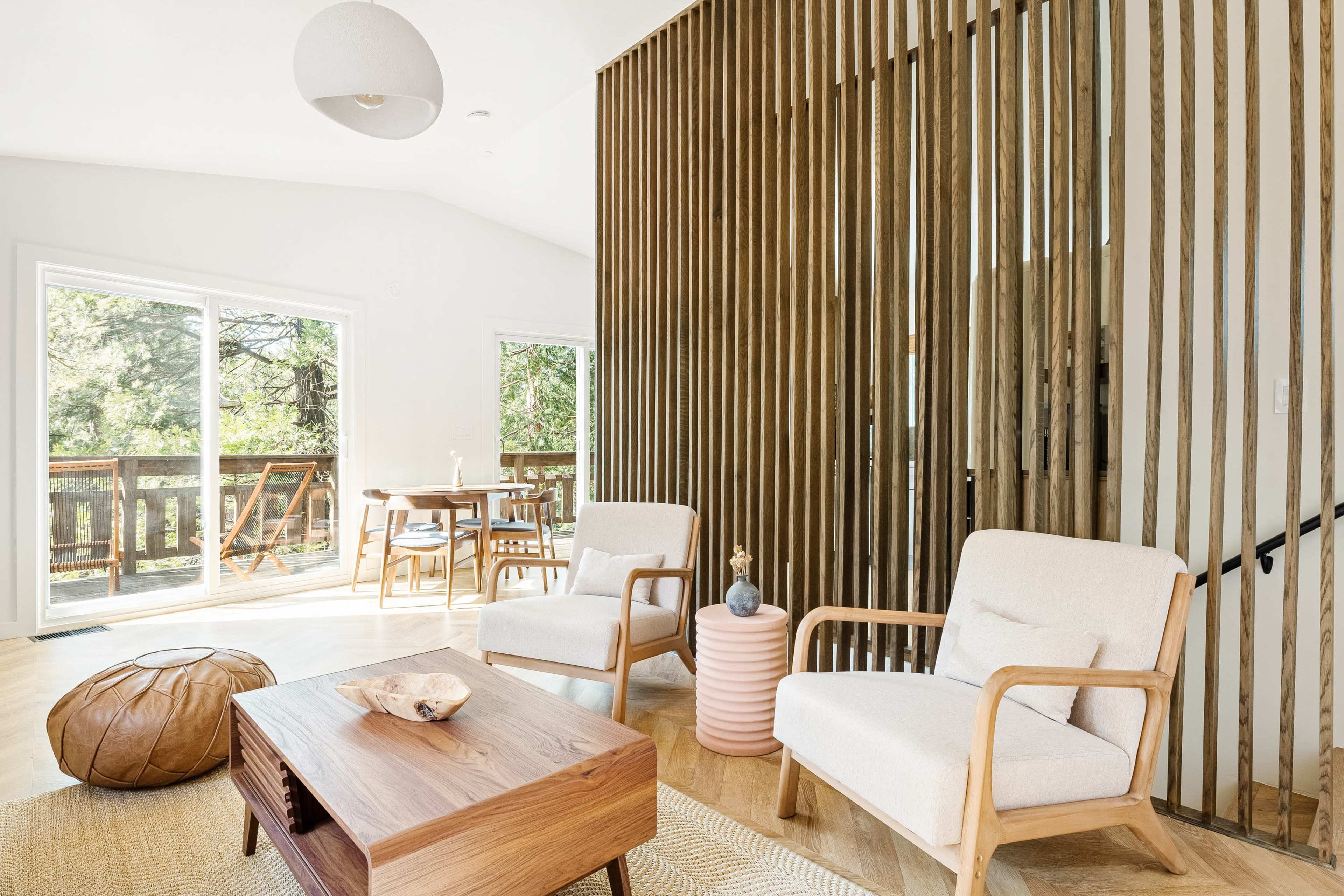 A contemporary living area featuring two armchairs, a wooden coffee table, and a pink side table, with large windows revealing a view of trees outside.