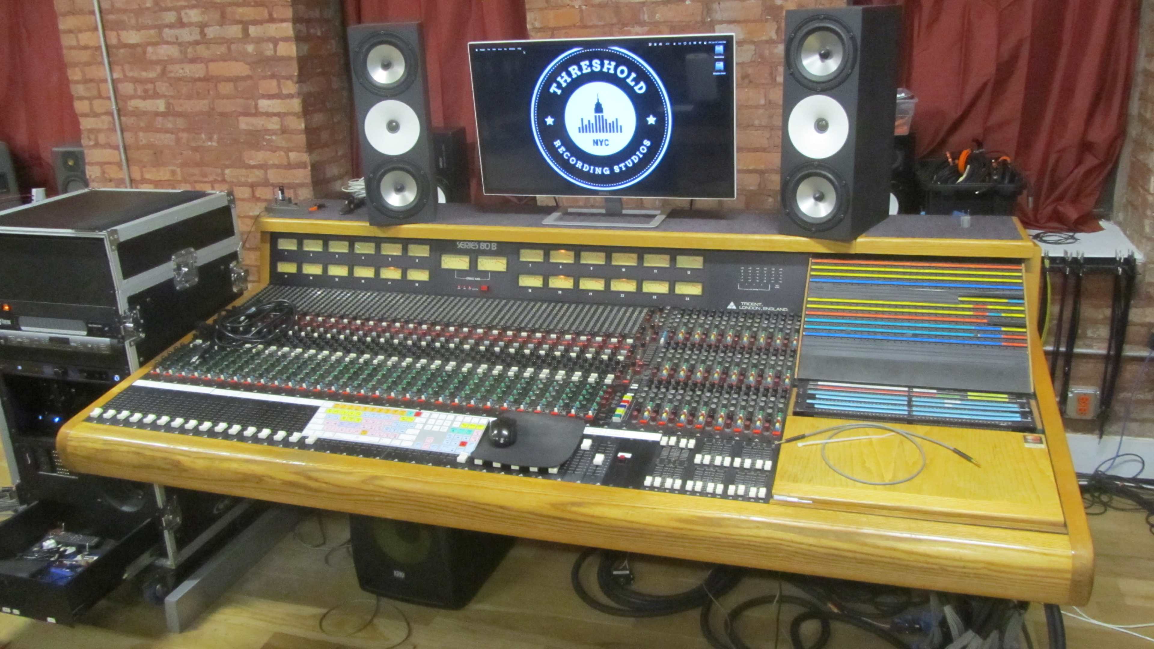 A large analog mixing console with colorful faders and a computer monitor displaying a logo is set up in a music studio.