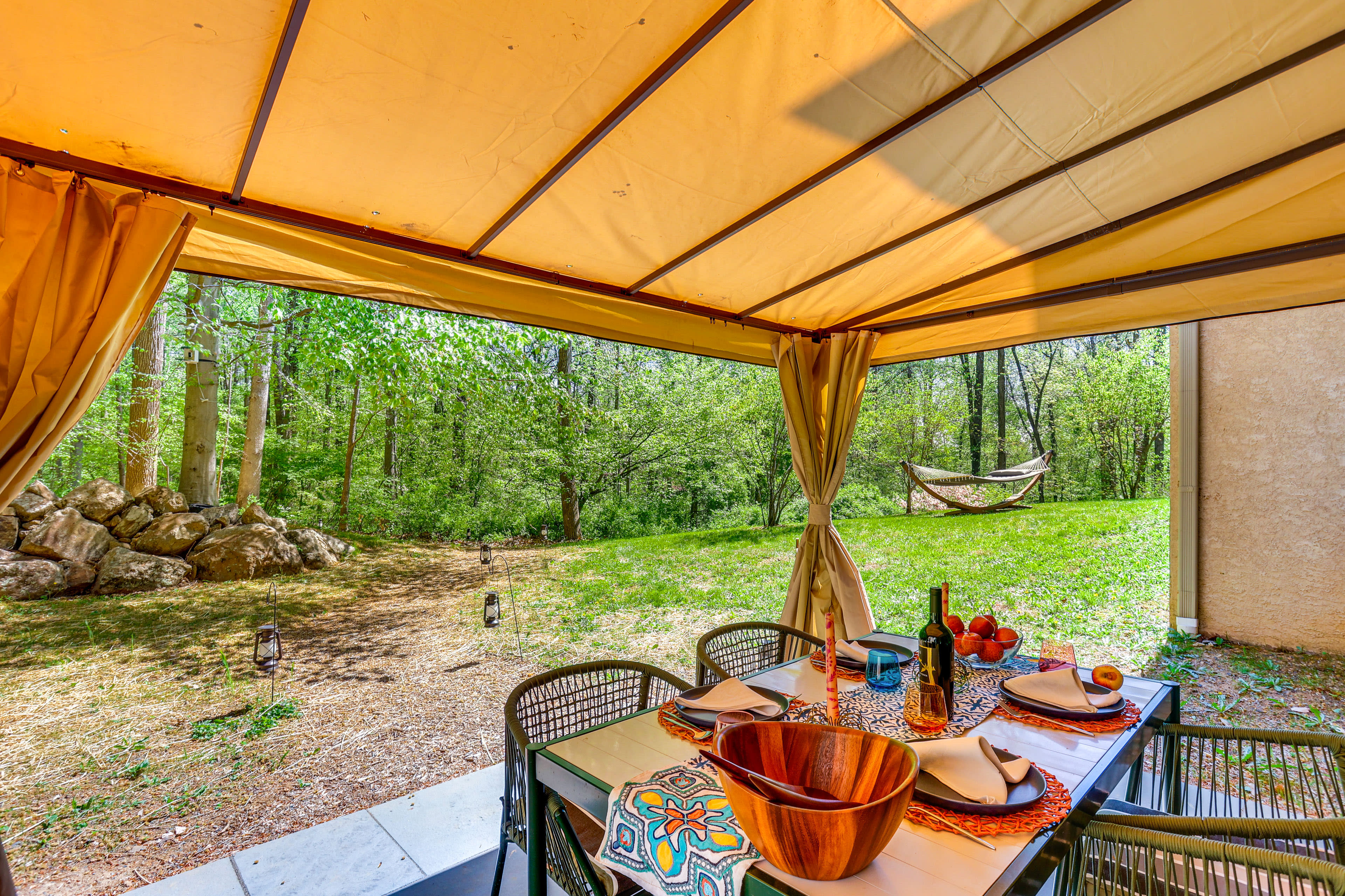 A dining table is set under a tent with a view of a grassy area featuring a hammock and trees in the background.