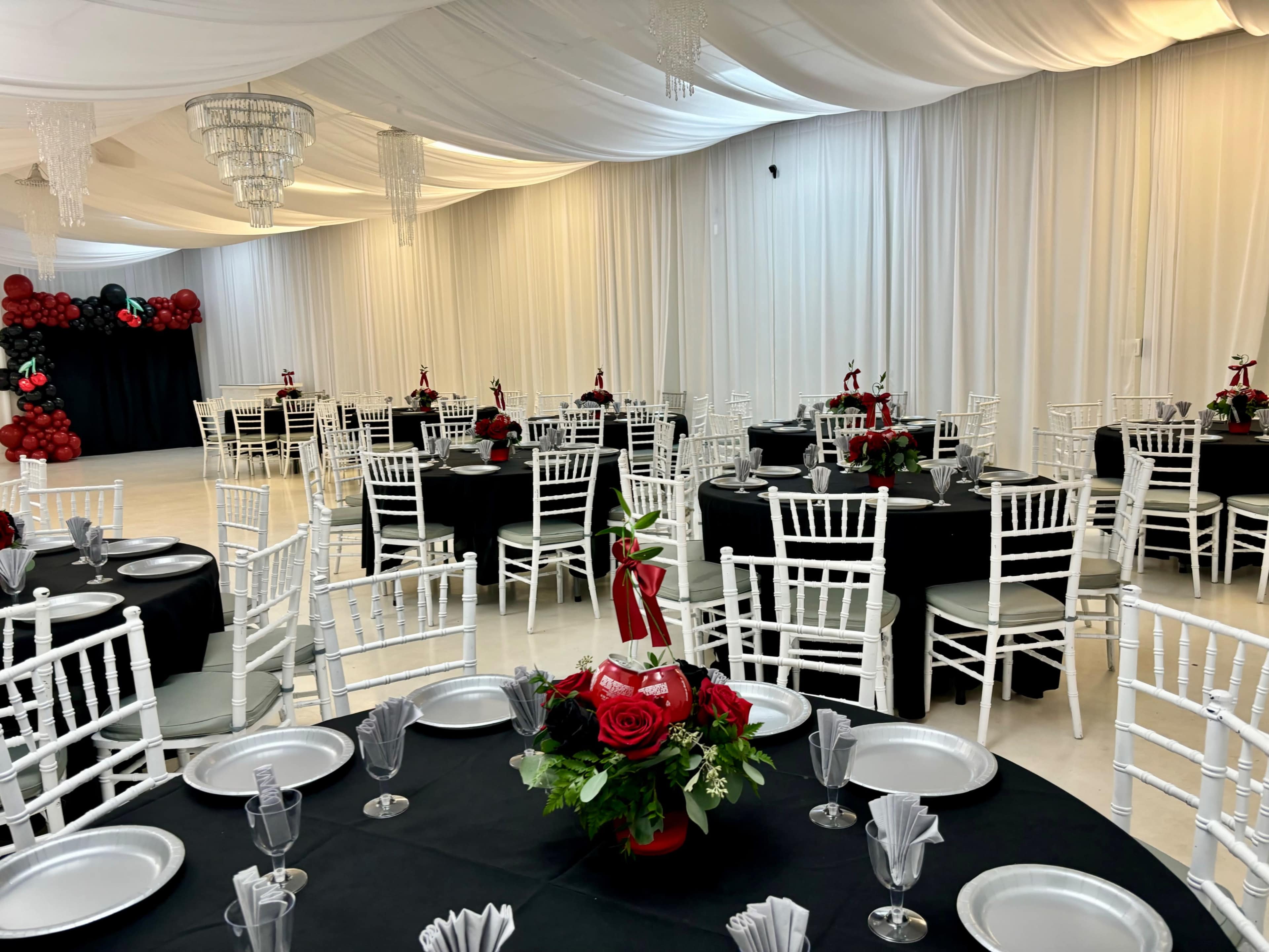 The image shows a banquet hall set up for an event, featuring round tables covered with black cloth and adorned with floral centerpieces, along with white chiavari chairs arranged around the tables.