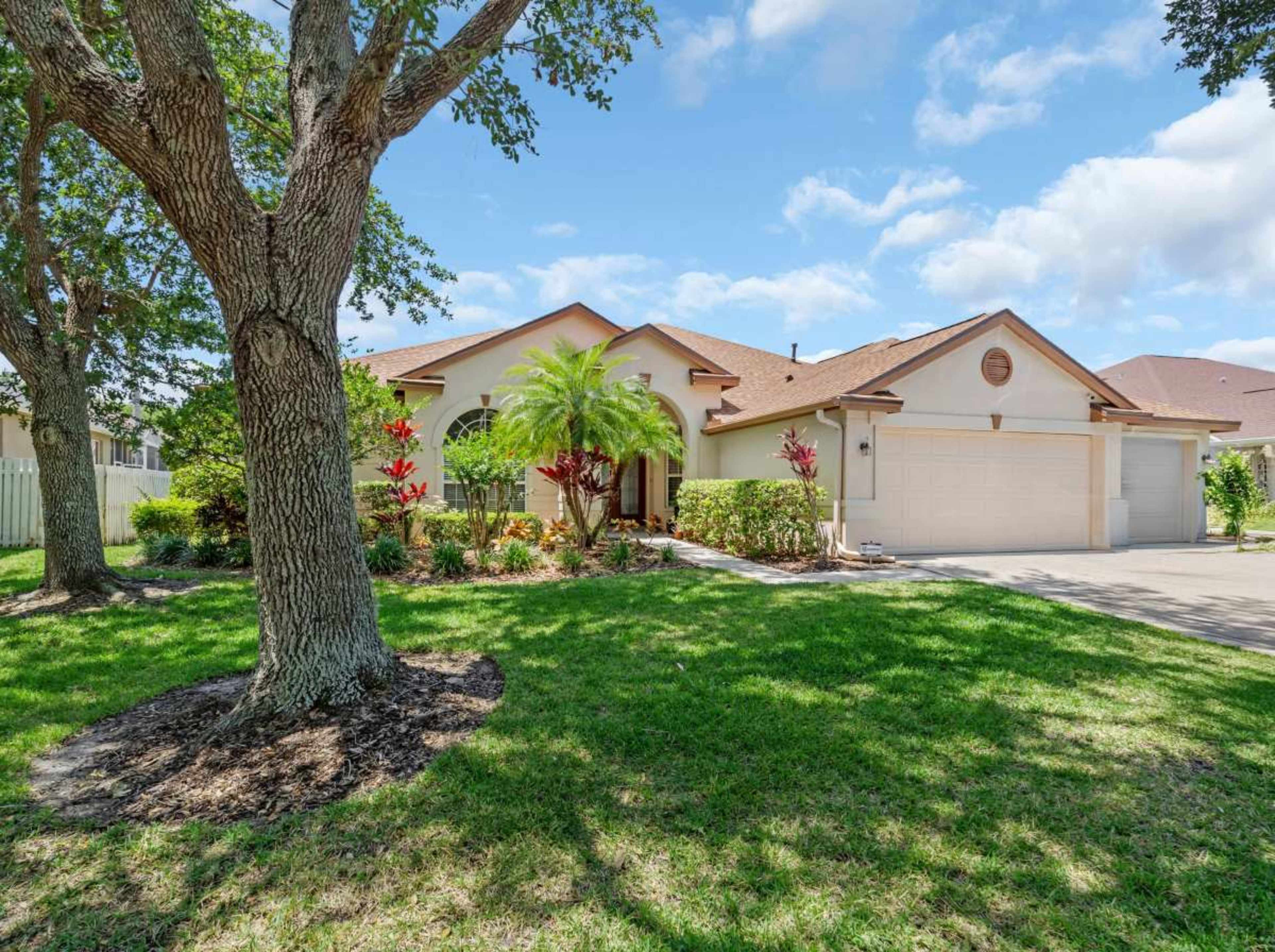 A single-story house with a landscaped front yard, surrounded by trees and colorful plants, features a two-car garage.