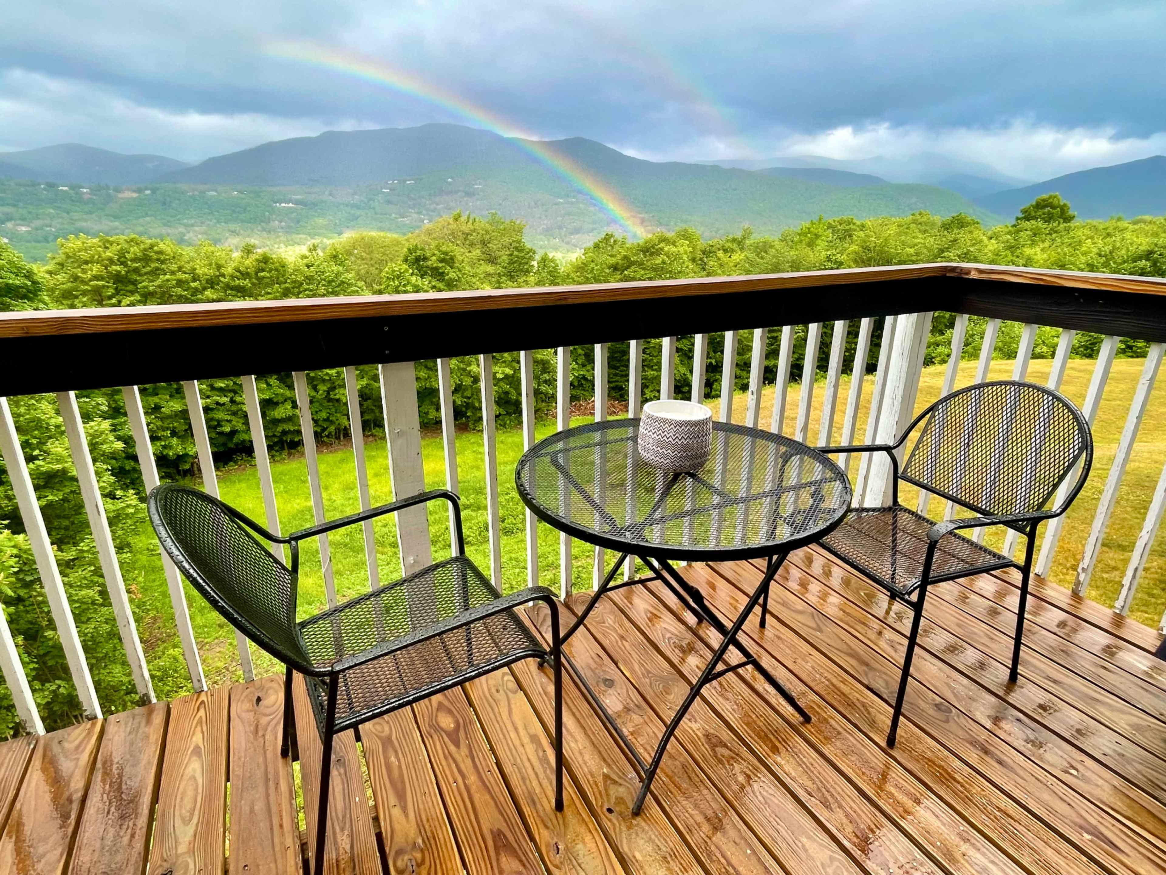 A wooden deck features two black metal chairs at a round table, with a rainbow visible over green mountains in the background.